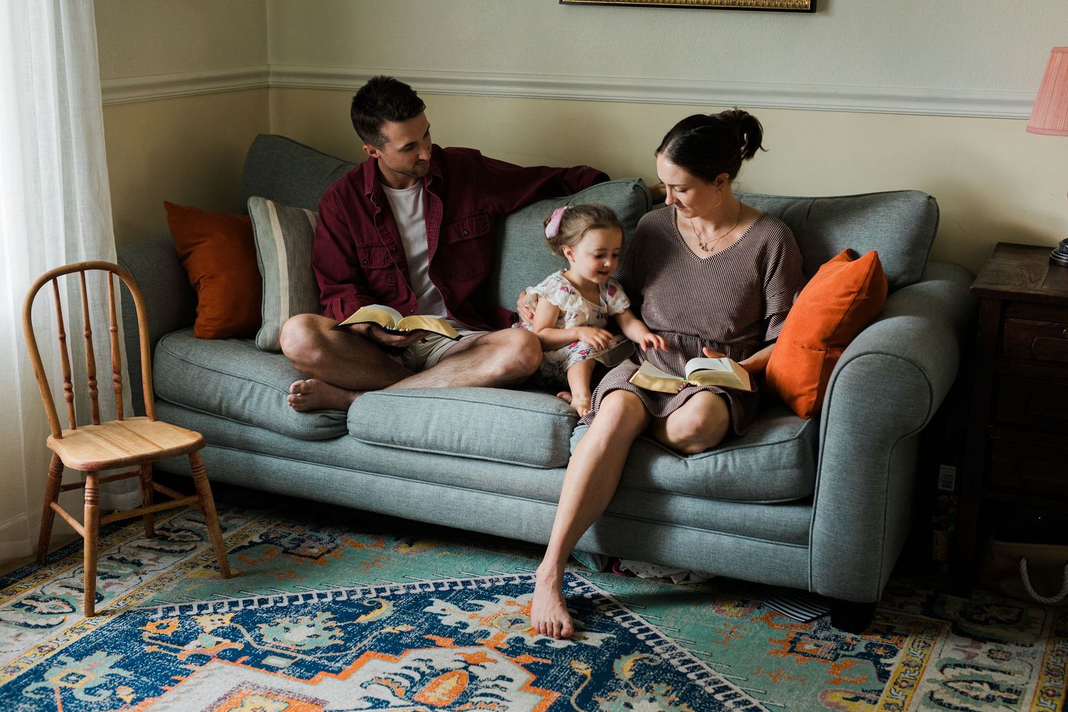 Family of three sitting on a gray sofa, parents reading books with baby in a cozy living room with blue rug.