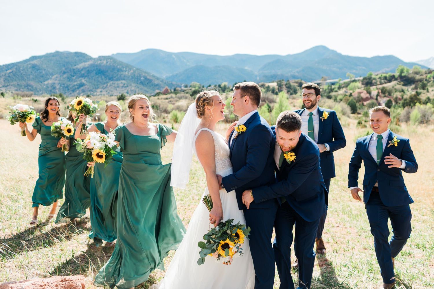 Wedding party in navy suits and green dresses with sunflowers celebrating in Colorado mountain field.