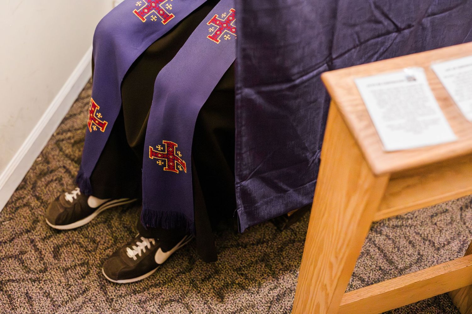Clergy member wearing purple religious stole with cross symbols and Nike sneakers seated beside a satin-draped surface.