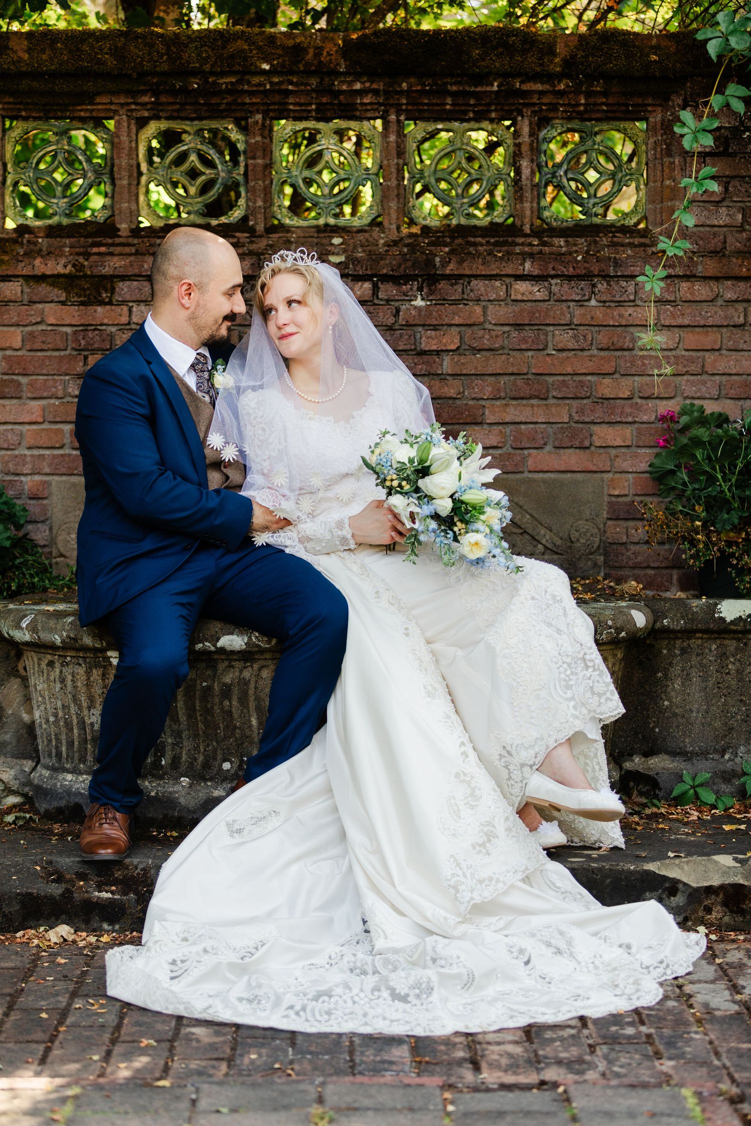 Bride and groom sit together on a stone bench in a garden, bride holding a white and blue bouquet.