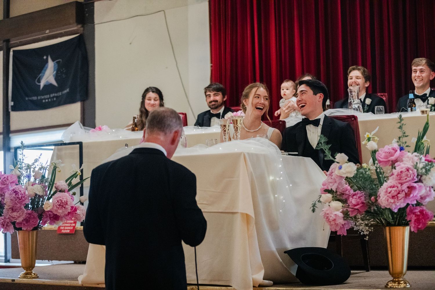 Wedding reception guests sit at round tables with pink floral centerpieces in a banquet hall with red curtains.