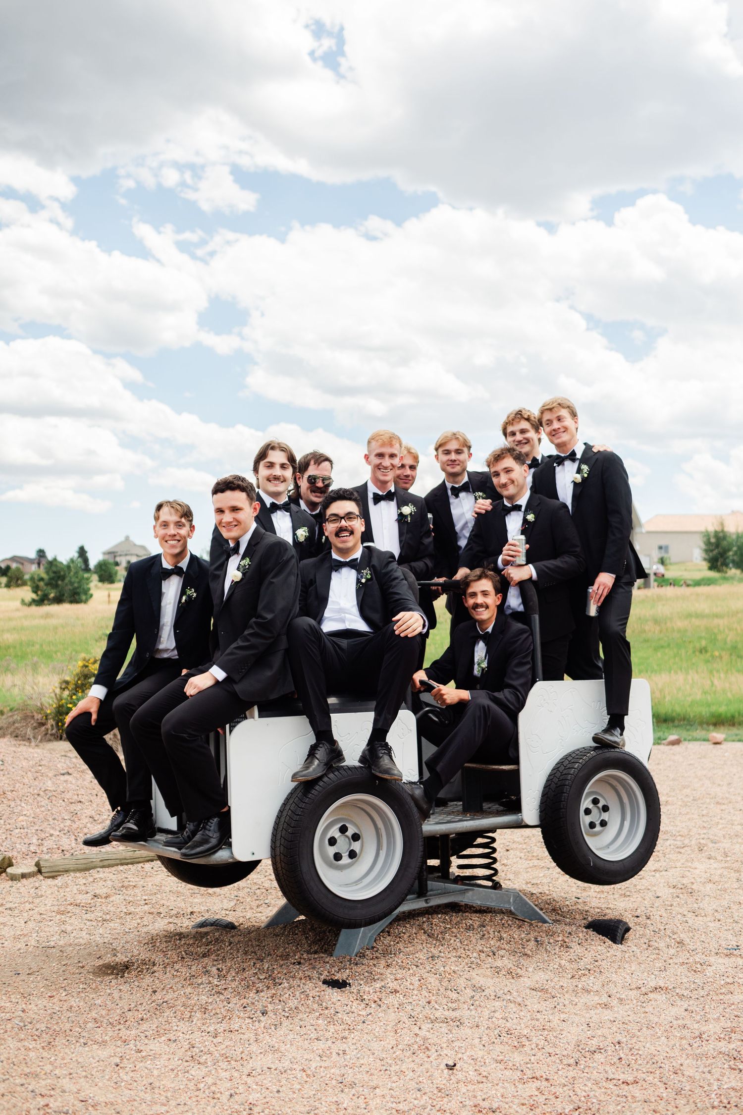 Groomsmen in black tuxedos pose playfully on a large white outdoor vehicle.