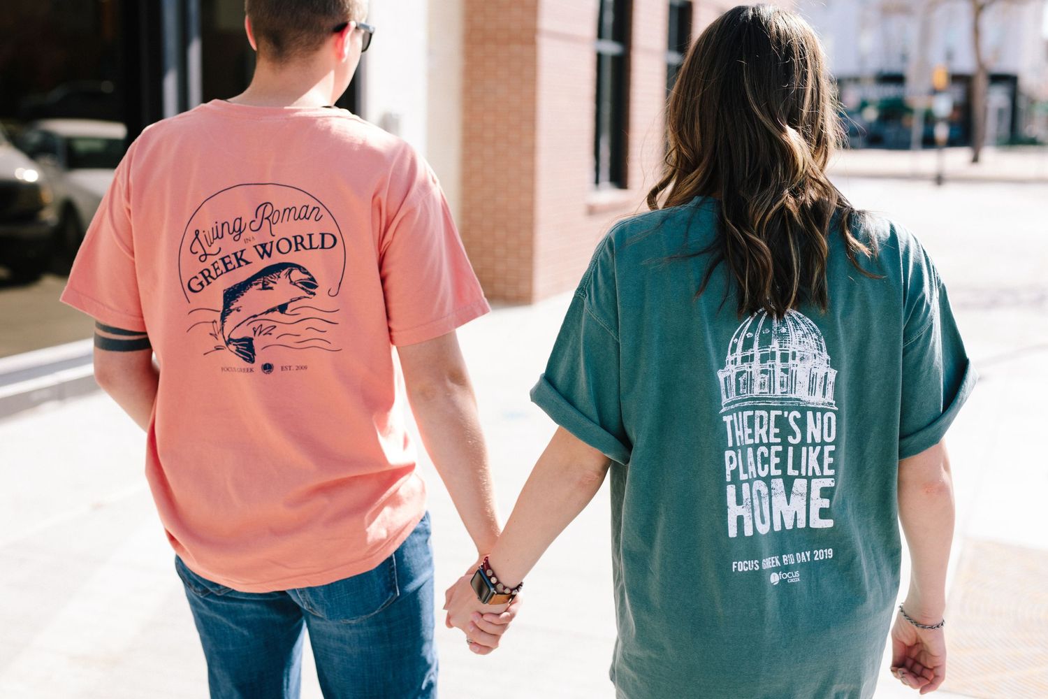 Two people wearing casual t-shirts with graphic designs hold hands while walking together outdoors in sunlight.