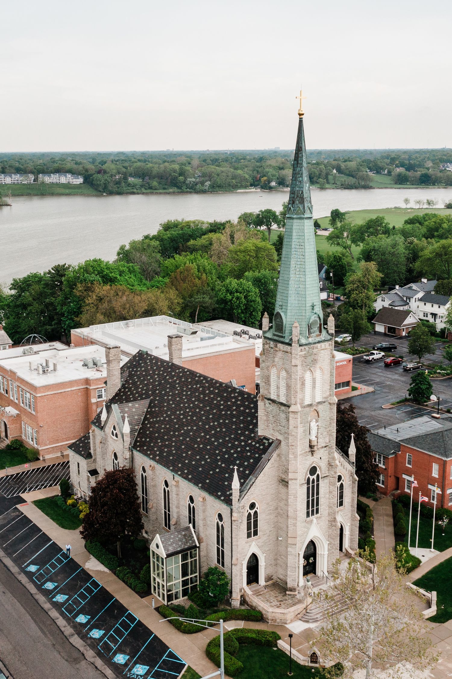Aerial view of a historic stone Gothic church with green copper steeple in a small riverside town surrounded by trees.