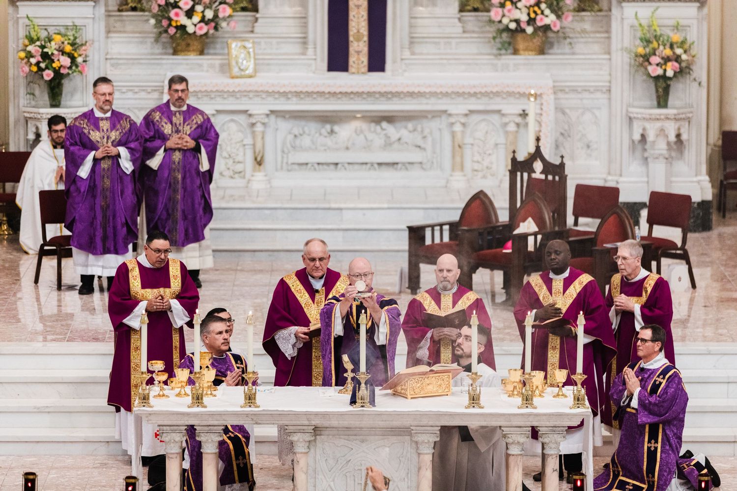 Catholic priests in purple vestments celebrating Mass at ornate altar with candles and flowers.