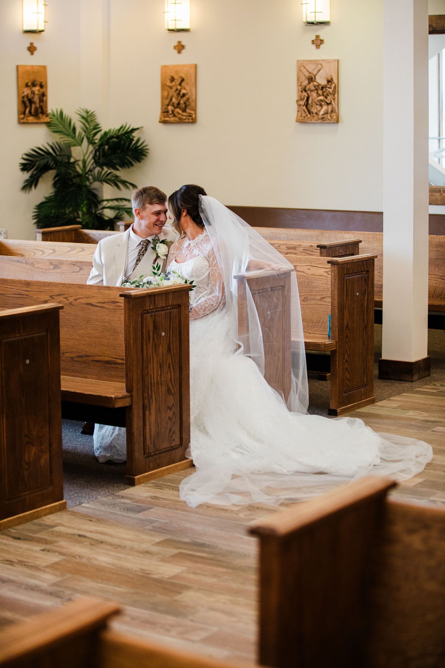 Bride and groom share an intimate moment seated in wooden church pews, bride wearing a flowing white gown and veil.