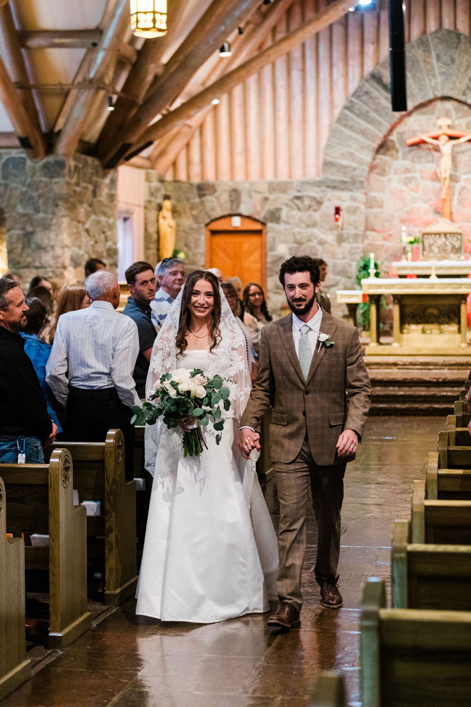 Bride and groom walk down church aisle after wedding ceremony, bride holding white floral bouquet in stone chapel.