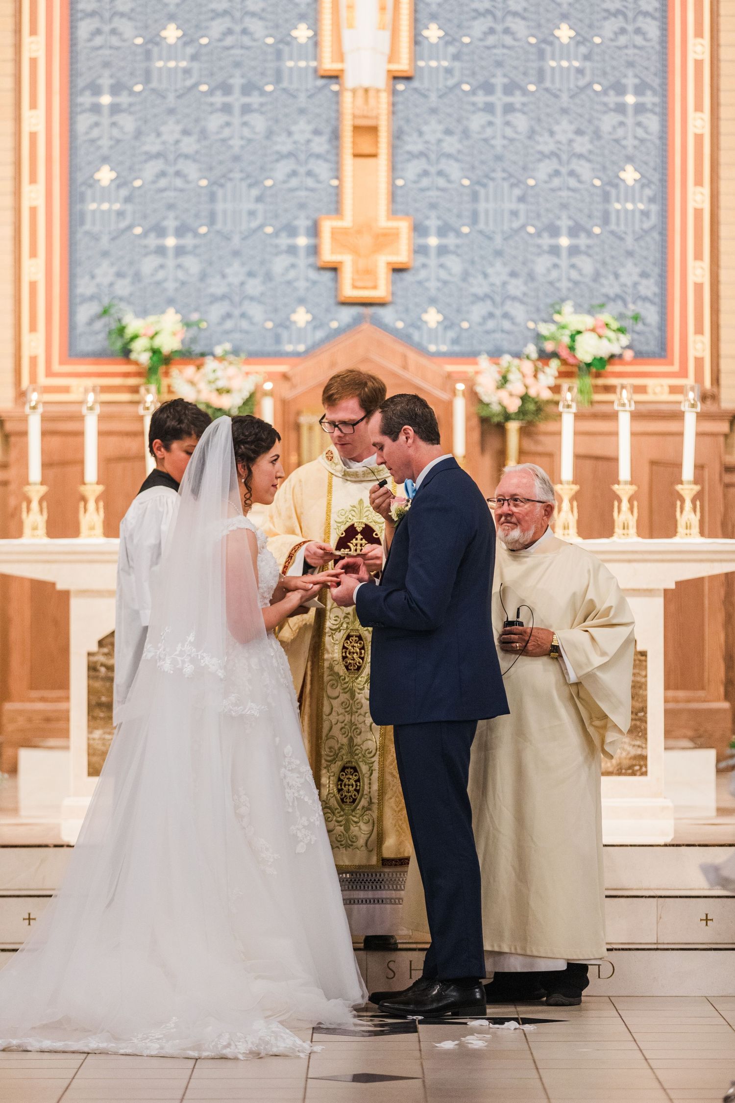 Bride and groom exchange rings at church altar with priest officiating Catholic wedding ceremony.