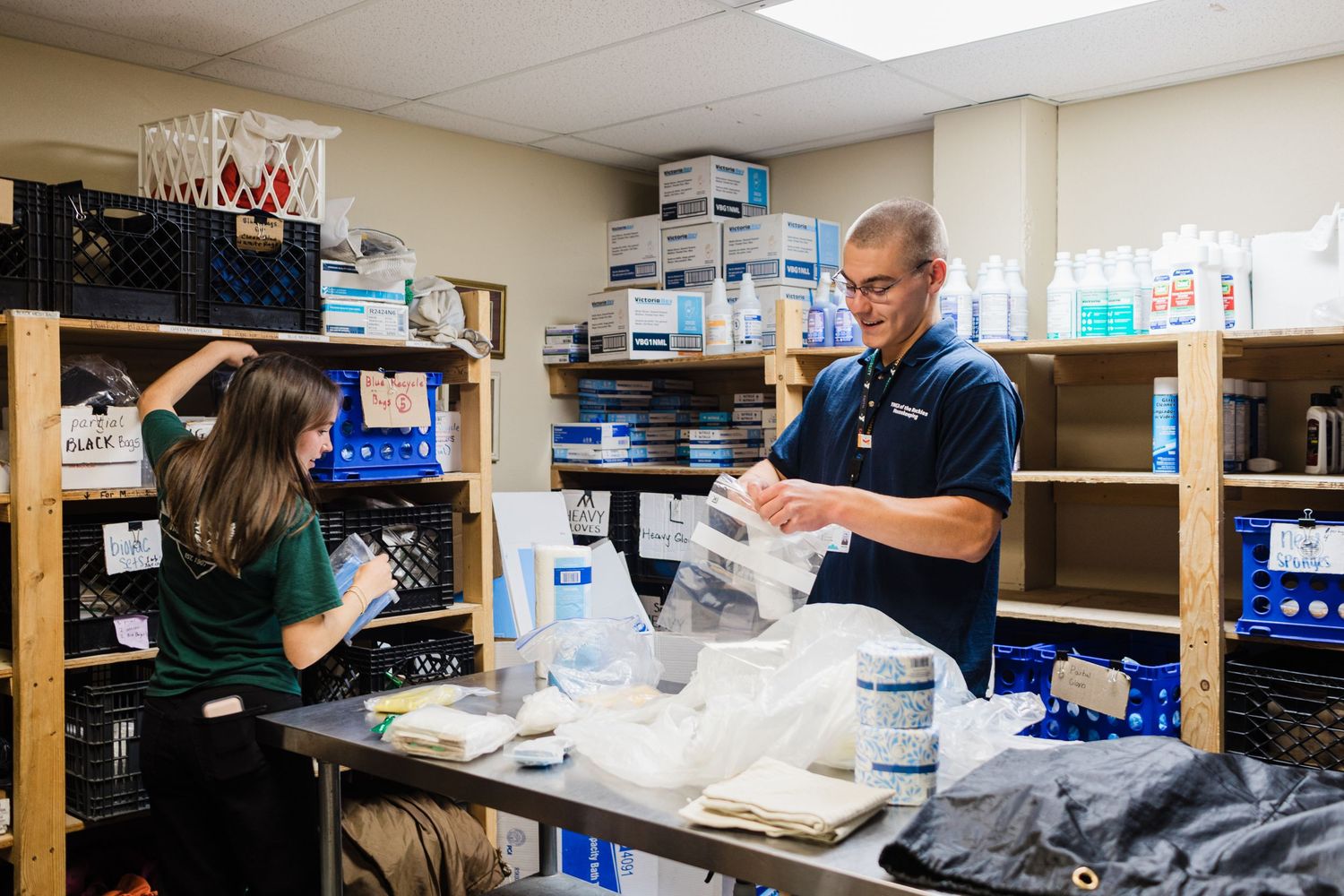 Two workers organizing medical supplies and packaging materials in a well-stocked storage room with shelves.