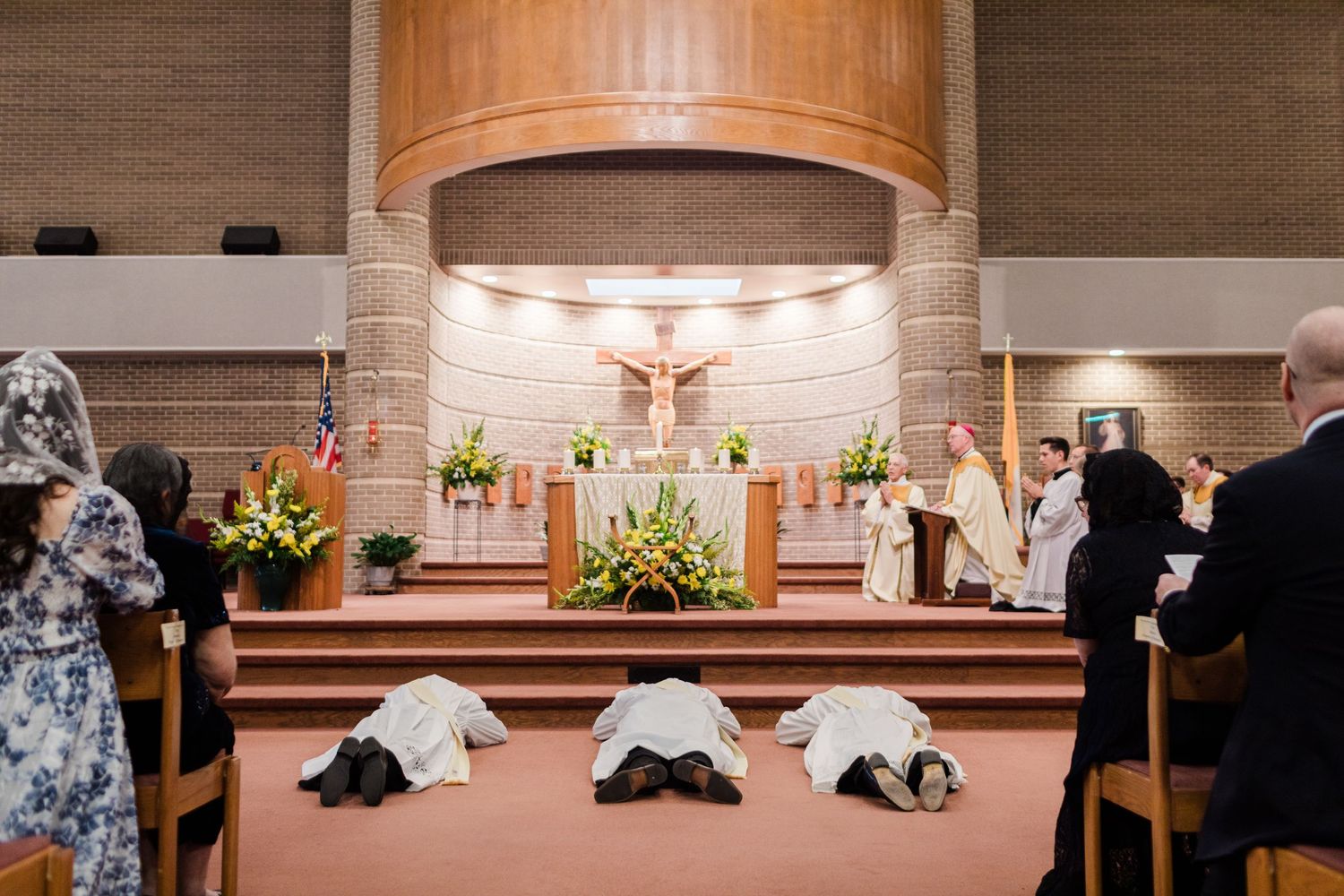 Catholic ordination ceremony with prostrate candidates before altar, bishop officiating, congregation standing in modern church.