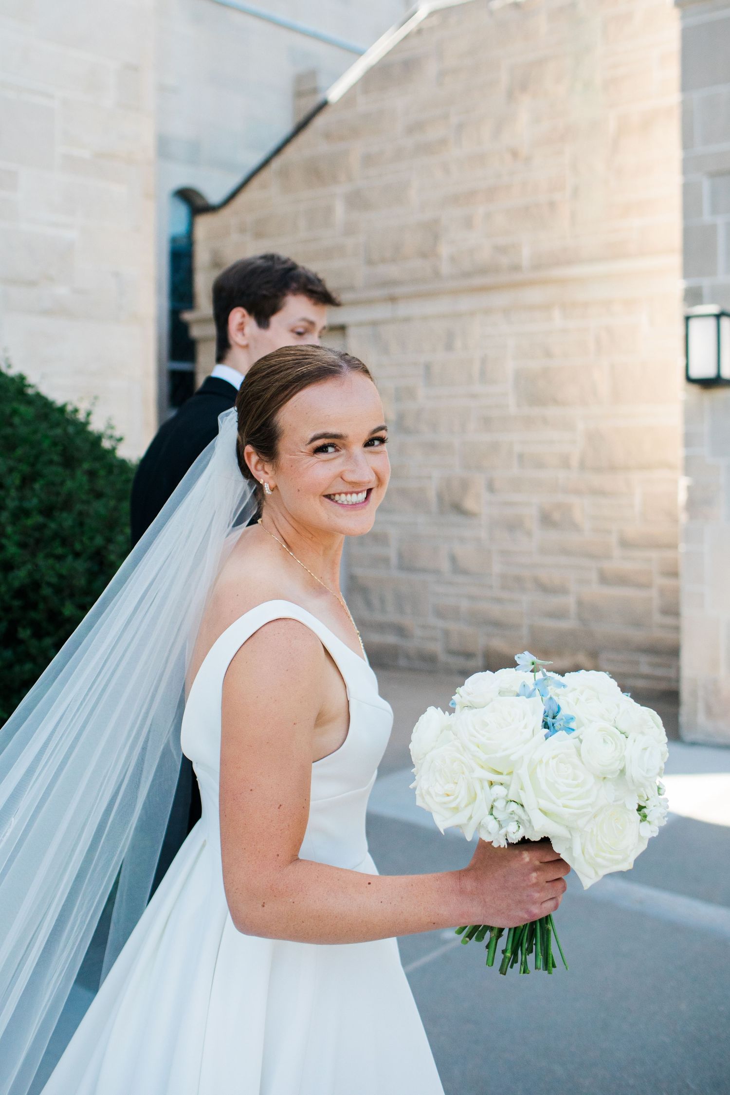 Smiling bride in white gown holds bouquet of white roses with blue accents, groom in black suit behind her.
