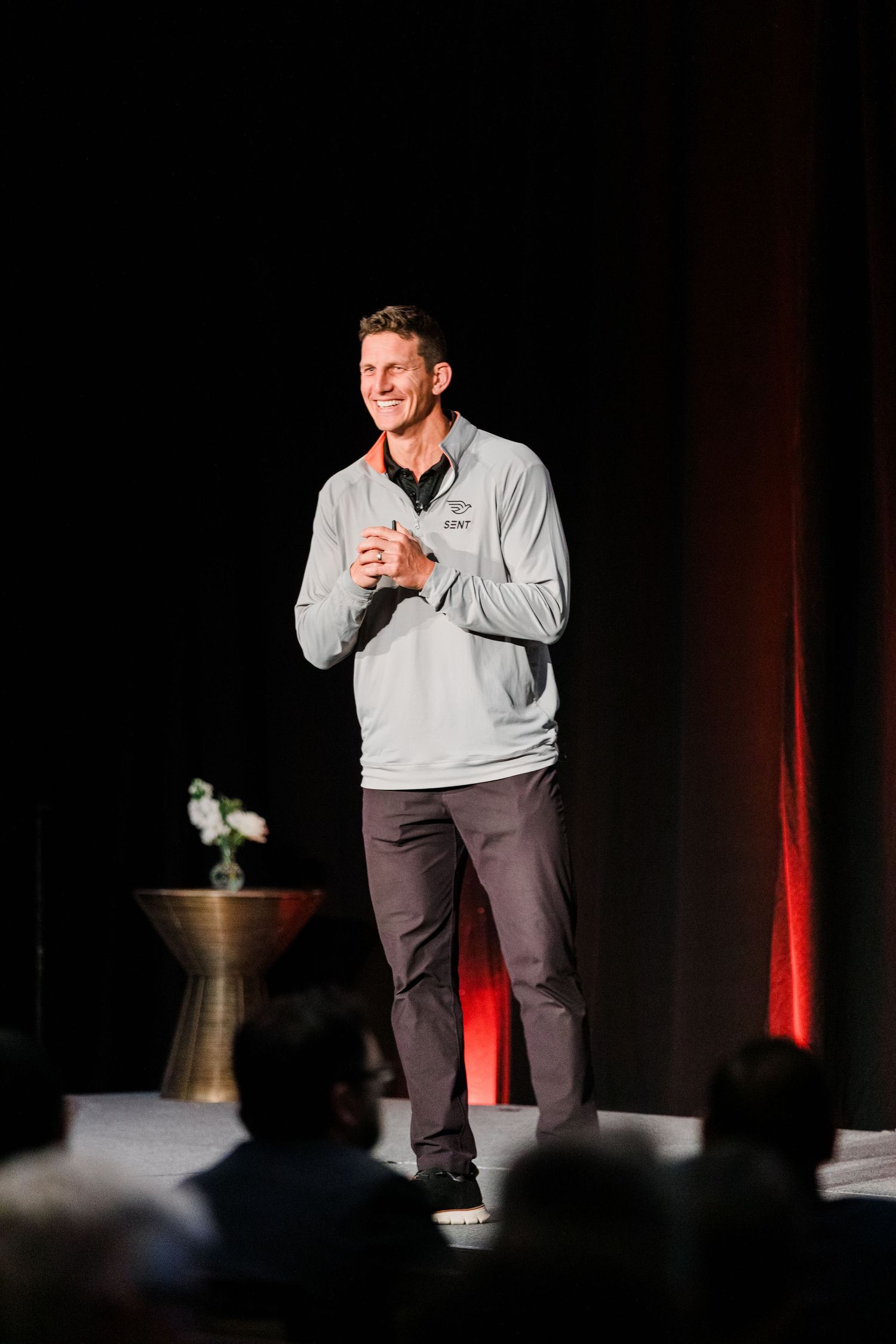 Speaker smiling on stage at Sent Summit 2026 wearing a gray pullover, holding a microphone in dramatic lighting.