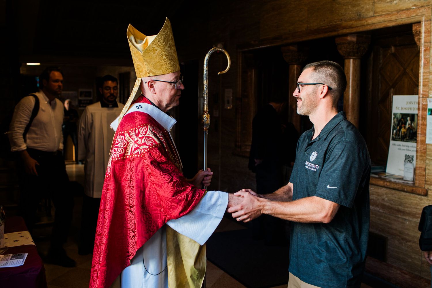 Bishop in red vestments shaking hands with a man in a green polo shirt inside a church.