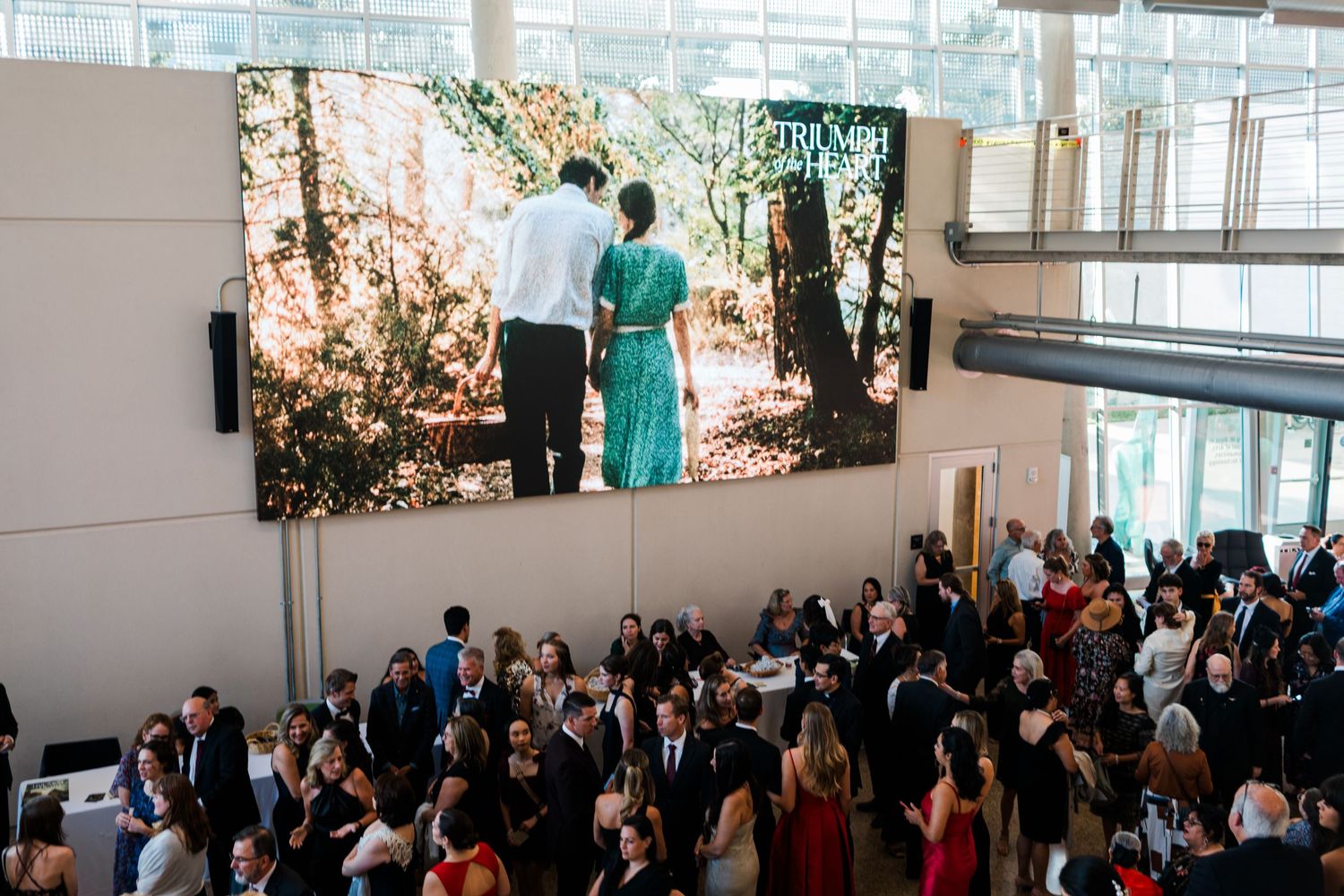 Large crowd gathers beneath a wall-mounted display showing a couple walking through a sunlit forest path.