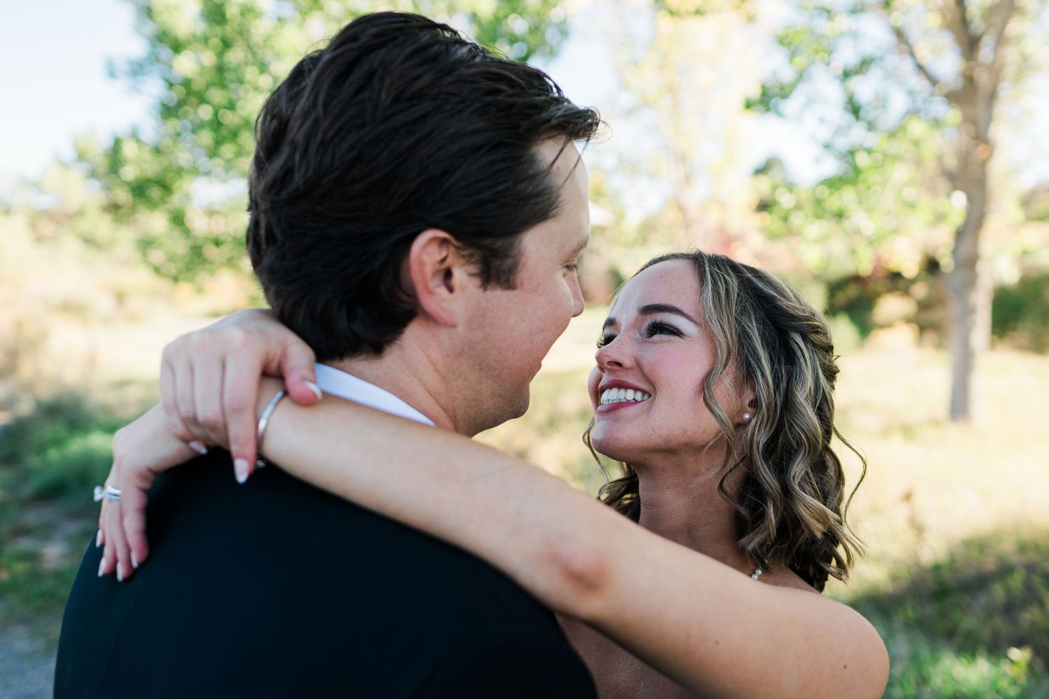 A smiling bride embraces her groom outdoors, gazing lovingly at each other during a romantic wedding photo session.