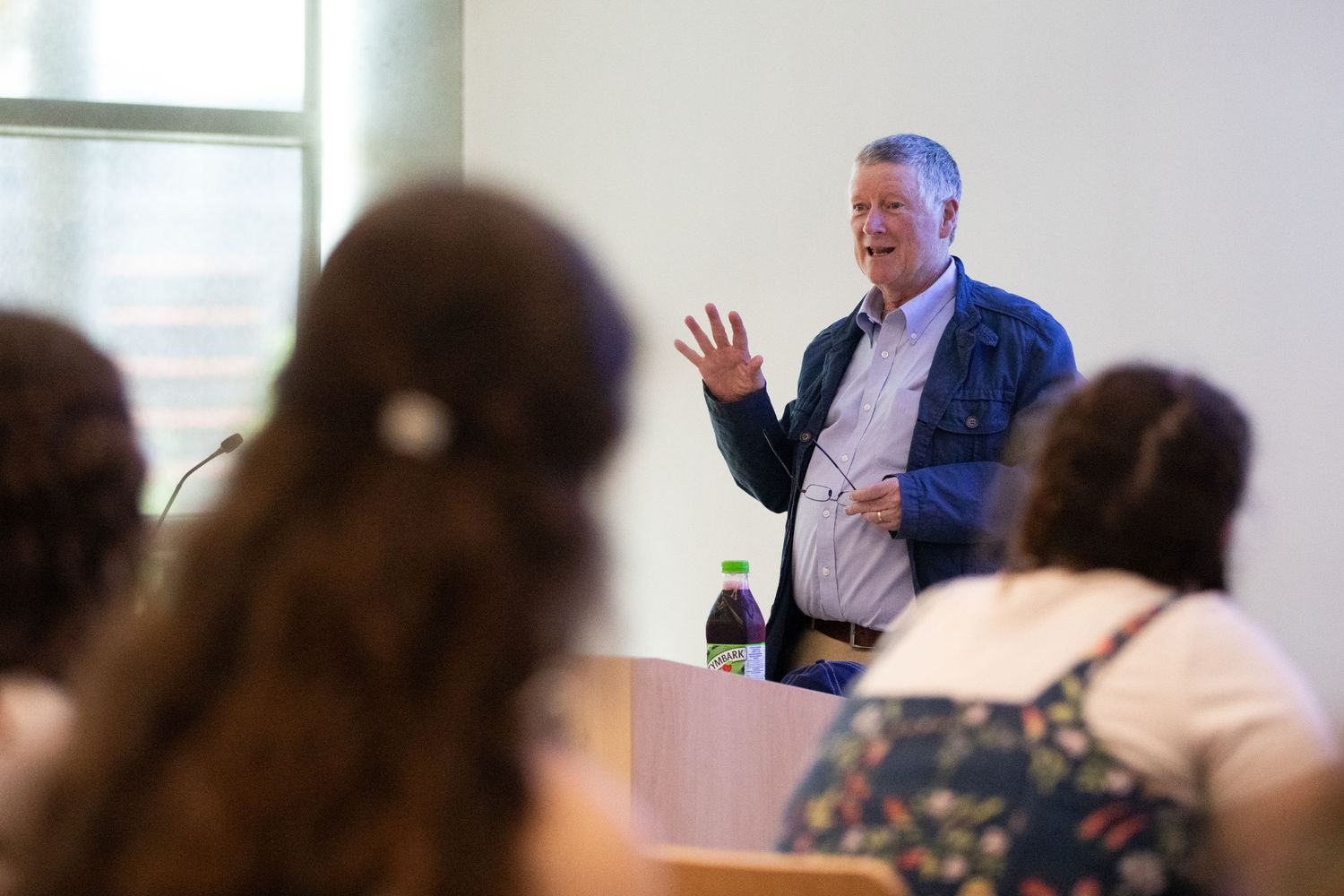 Professor gestures while speaking to students seated in a university lecture hall classroom setting.