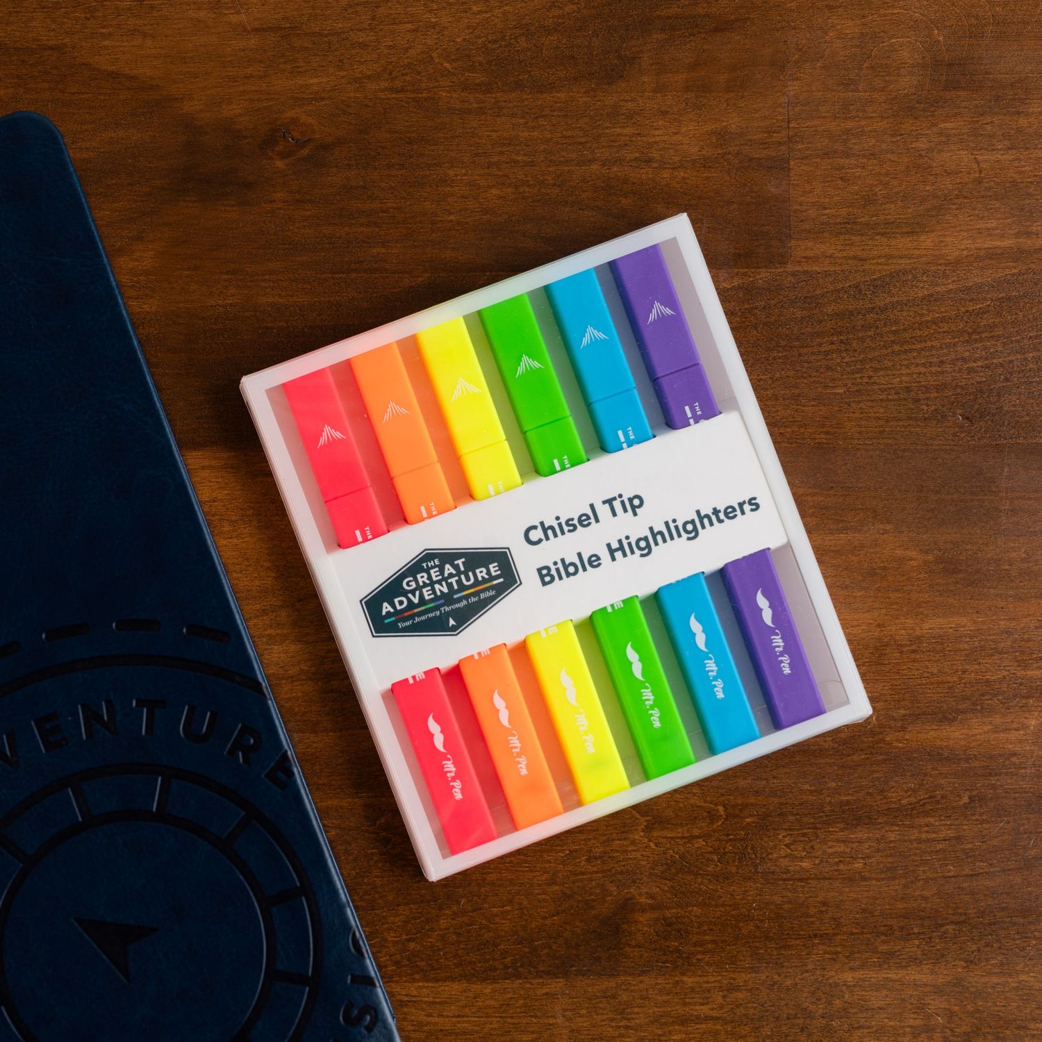 Rainbow colored chisel tip highlighter markers displayed in packaging on a wooden surface.