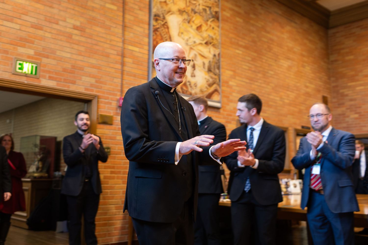 Catholic bishop in black vestments receives applause from attendees in formal attire at an indoor ceremony.
