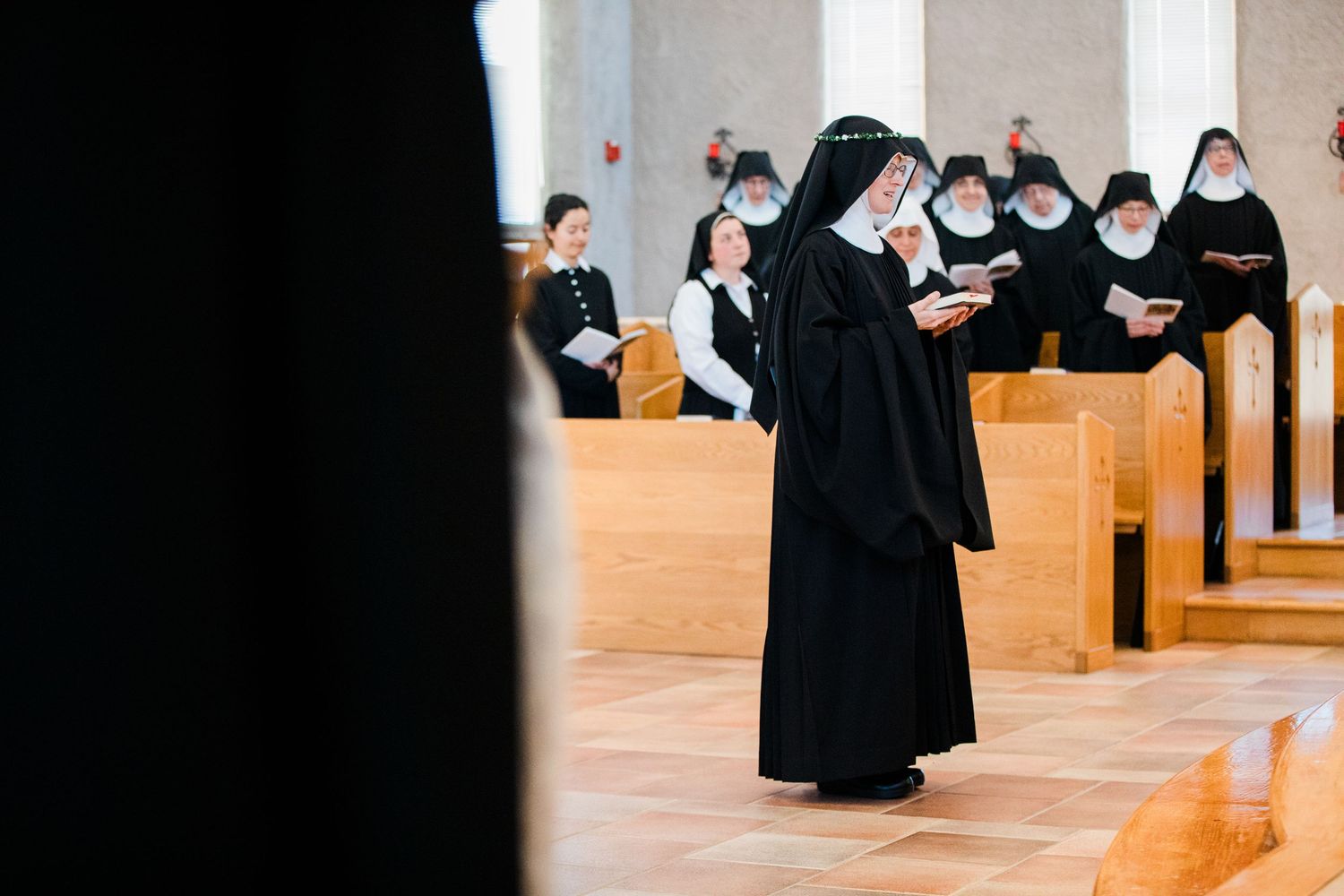 Catholic nuns in traditional black habits pray and sing during a religious ceremony inside a modern chapel.