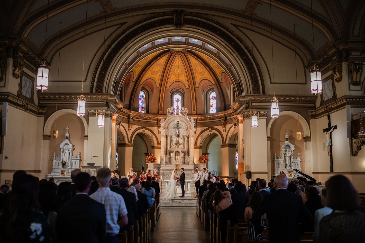Wedding ceremony inside a grand Catholic church with ornate altar, stained glass windows, and guests seated in wooden pews.