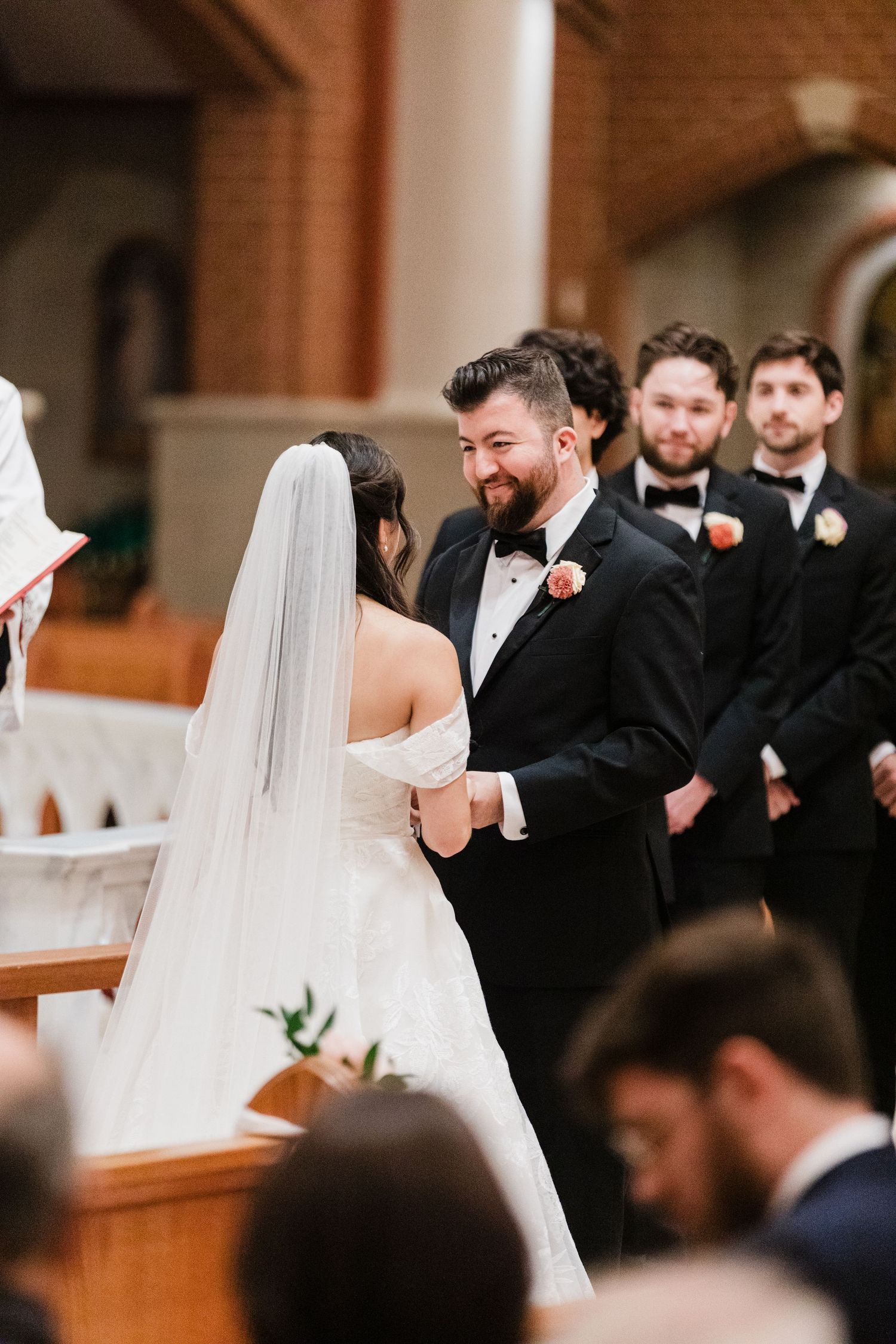 Groom smiling at bride during wedding ceremony at church altar, with groomsmen in black tuxedos standing behind.