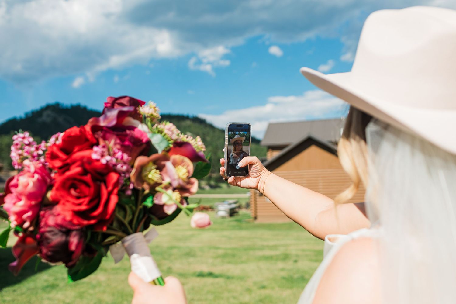 Woman in white hat photographing a vibrant red and pink bridal bouquet outdoors.