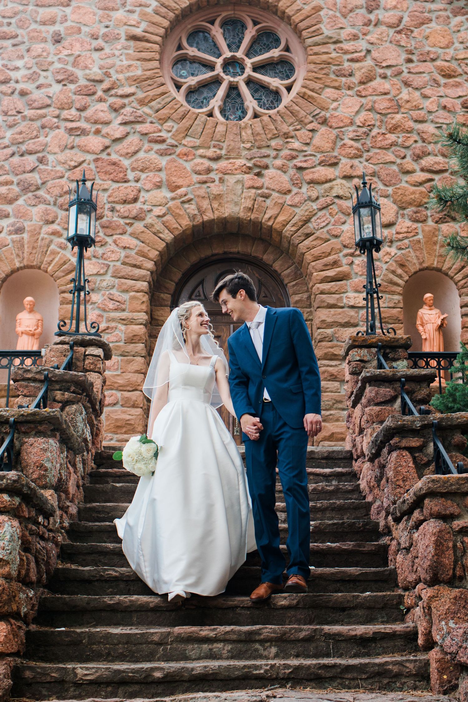 Bride and groom holding hands on stone church steps, bride in white gown with veil, groom in navy suit.