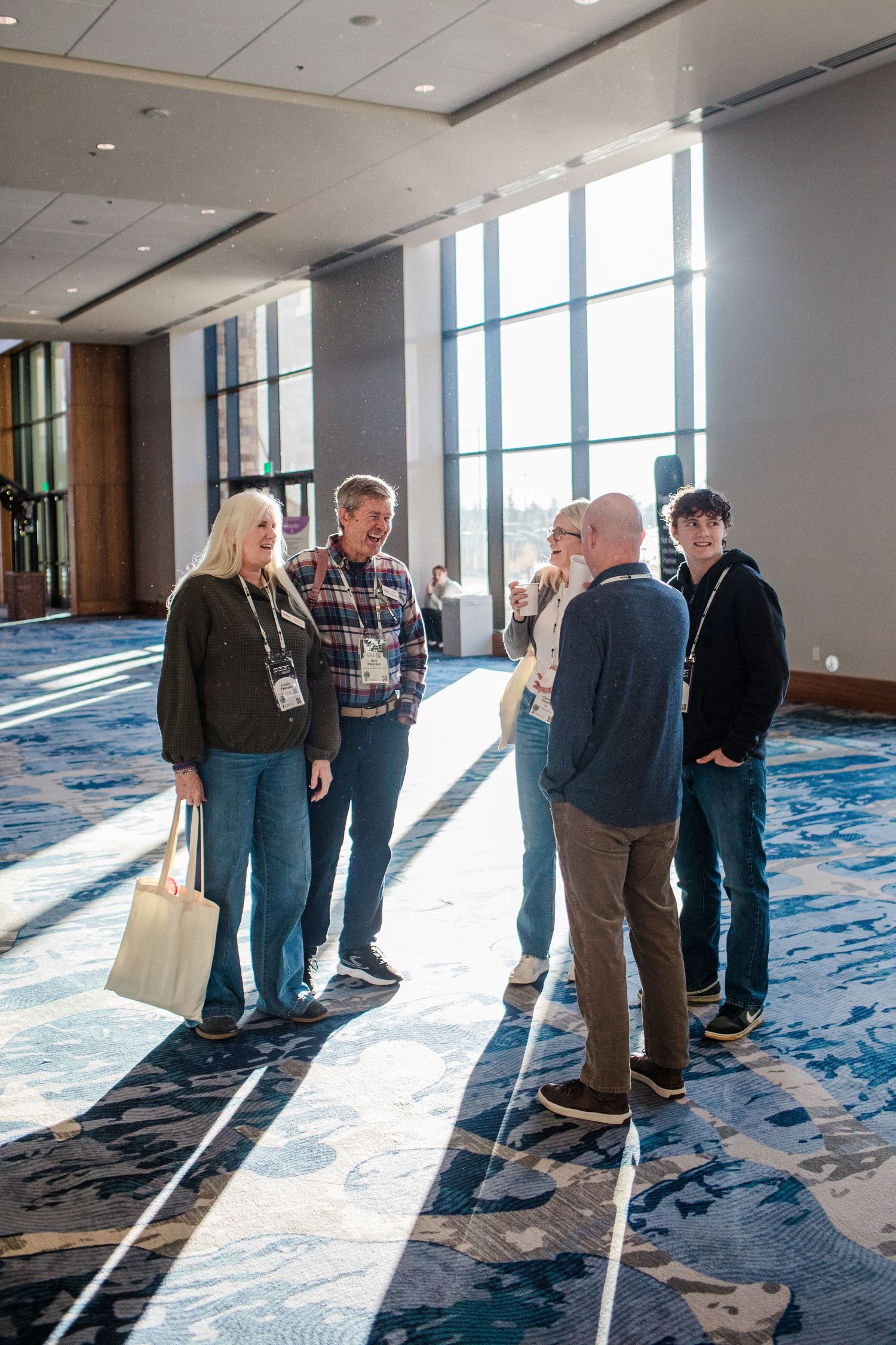People gathered in conversation near large windows in a modern conference center with blue carpeting.