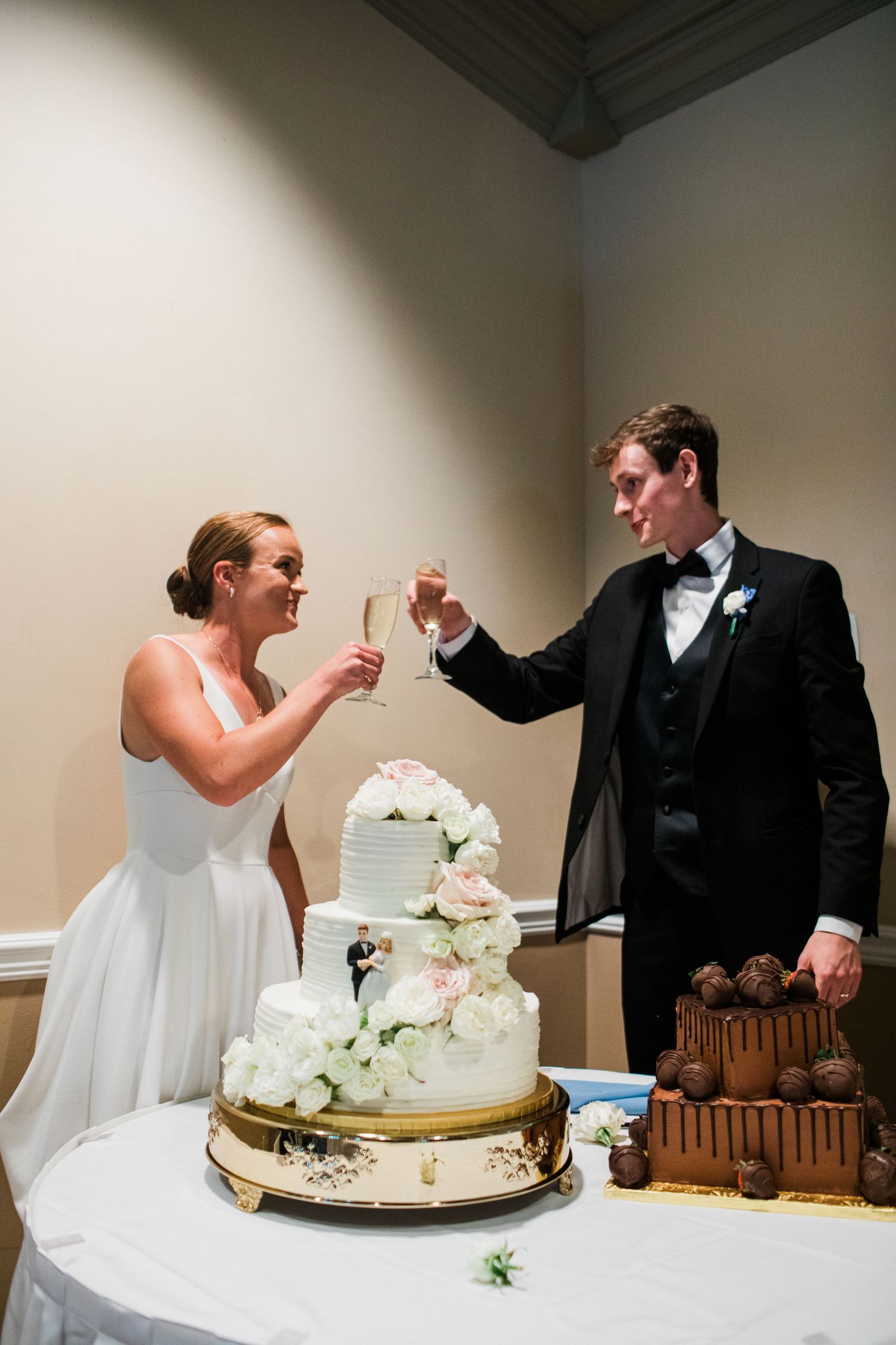 Bride and groom toasting with champagne beside a white floral wedding cake at their elegant reception.