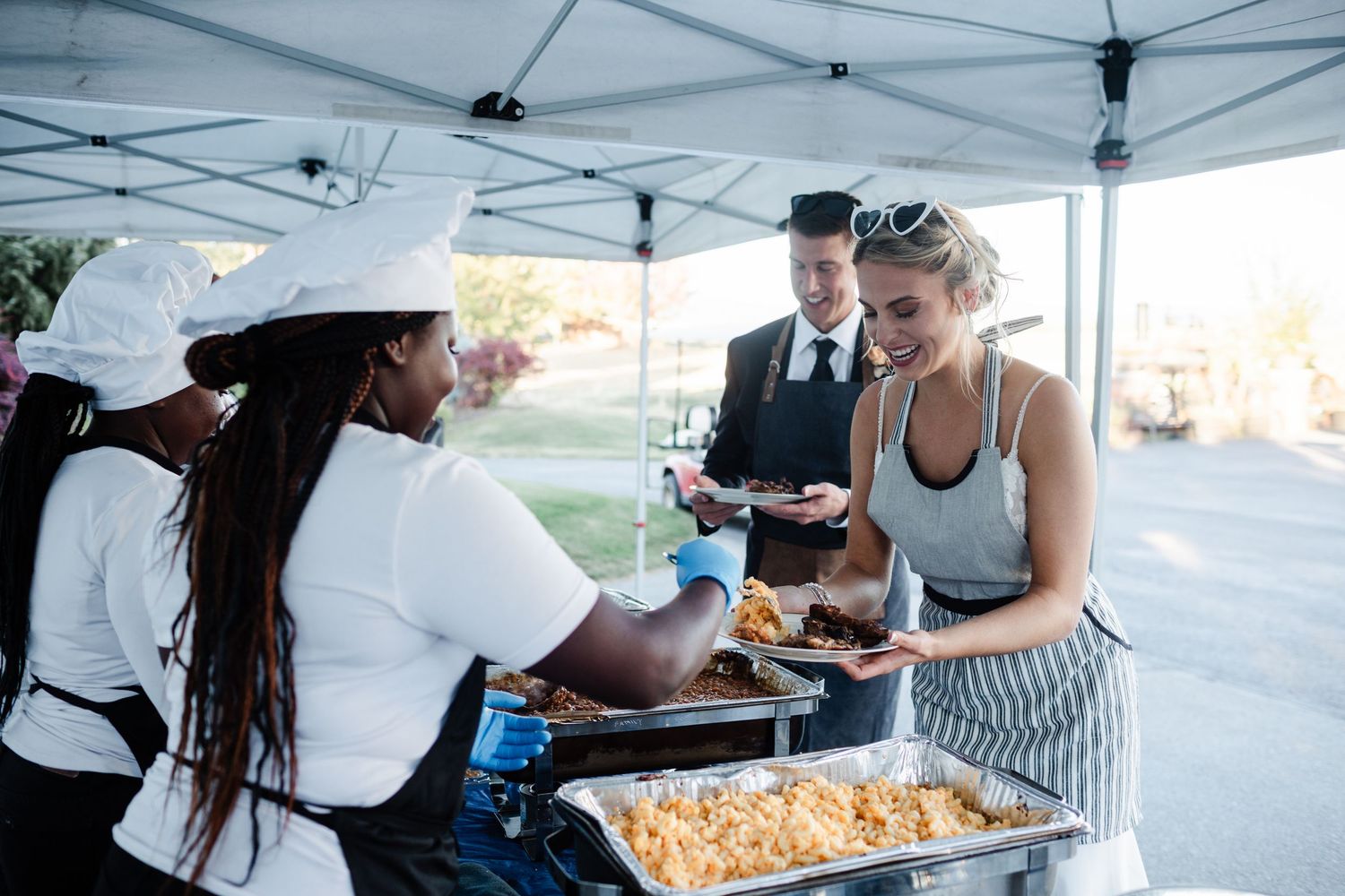 Outdoor food service event with staff serving buffet dishes to smiling guests under a canopy tent.