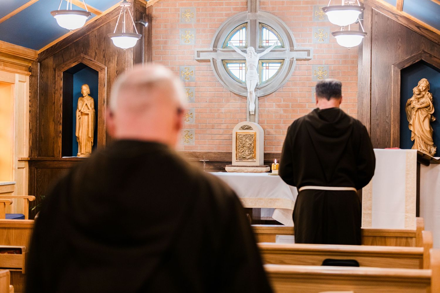 Two Franciscan monks in brown habits pray before a wooden cross altar inside a Catholic chapel.