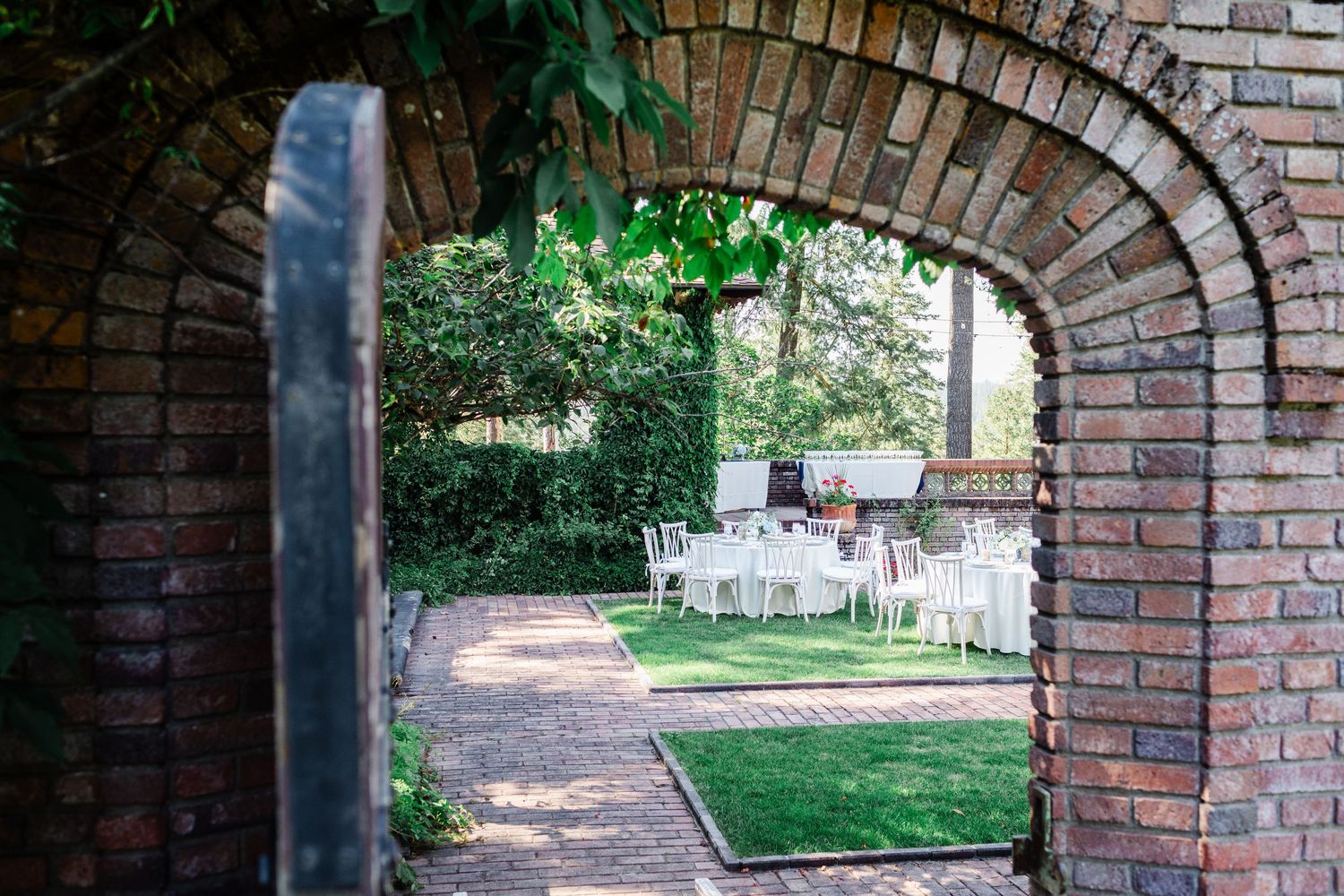Brick archway frames an elegant outdoor wedding reception with white tables and lush garden beyond.