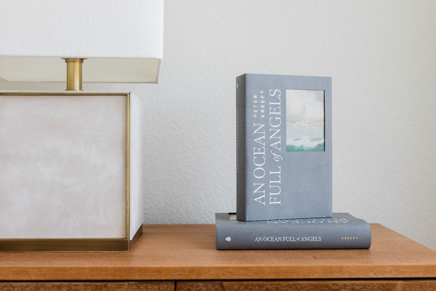 Two copies of 'An Ocean Full of Angels' book stacked on a wooden nightstand beside a modern lamp.