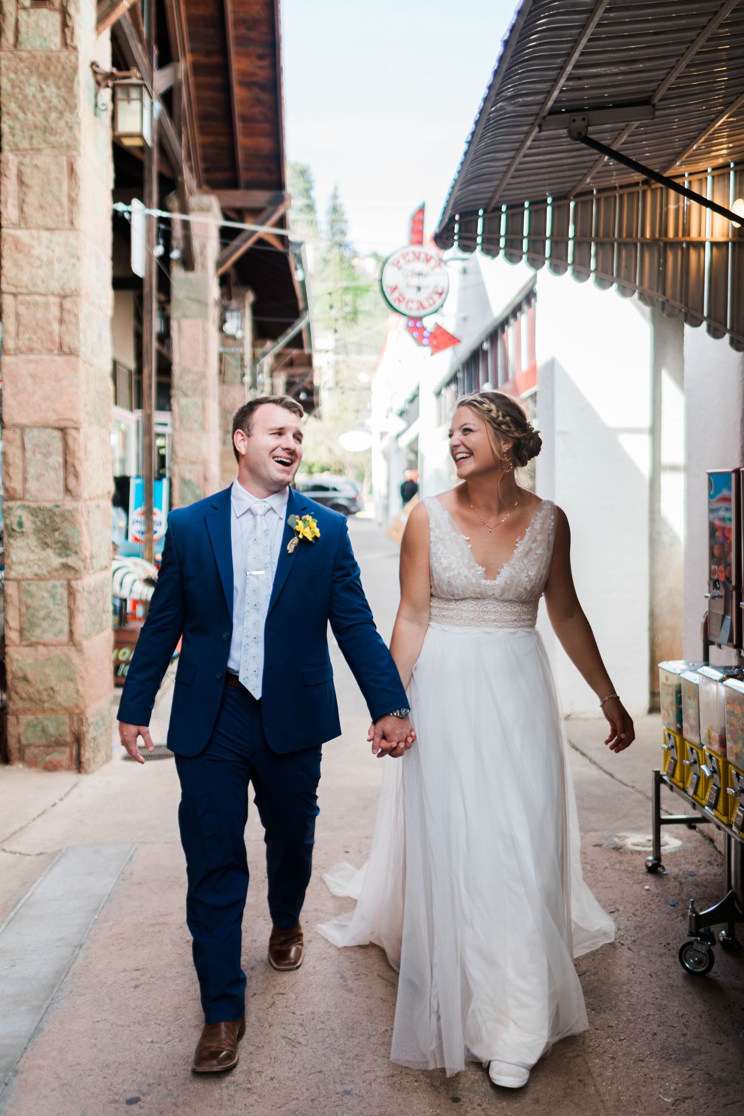 Smiling bride in white gown and groom in navy suit walk hand in hand through a charming Colorado street.