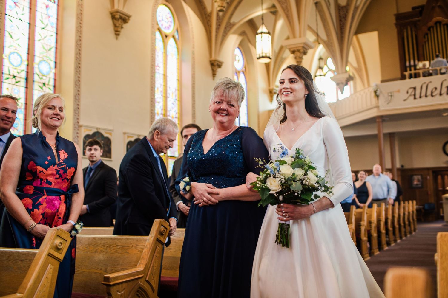 Bride walks down church aisle with smiling woman in blue dress, holding white bouquet during wedding ceremony.