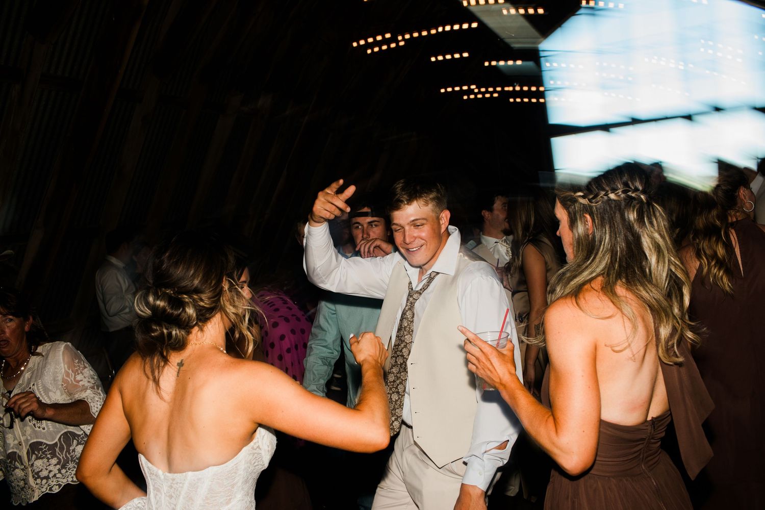 Guests dancing joyfully at a rustic wedding reception in a dimly lit barn venue.