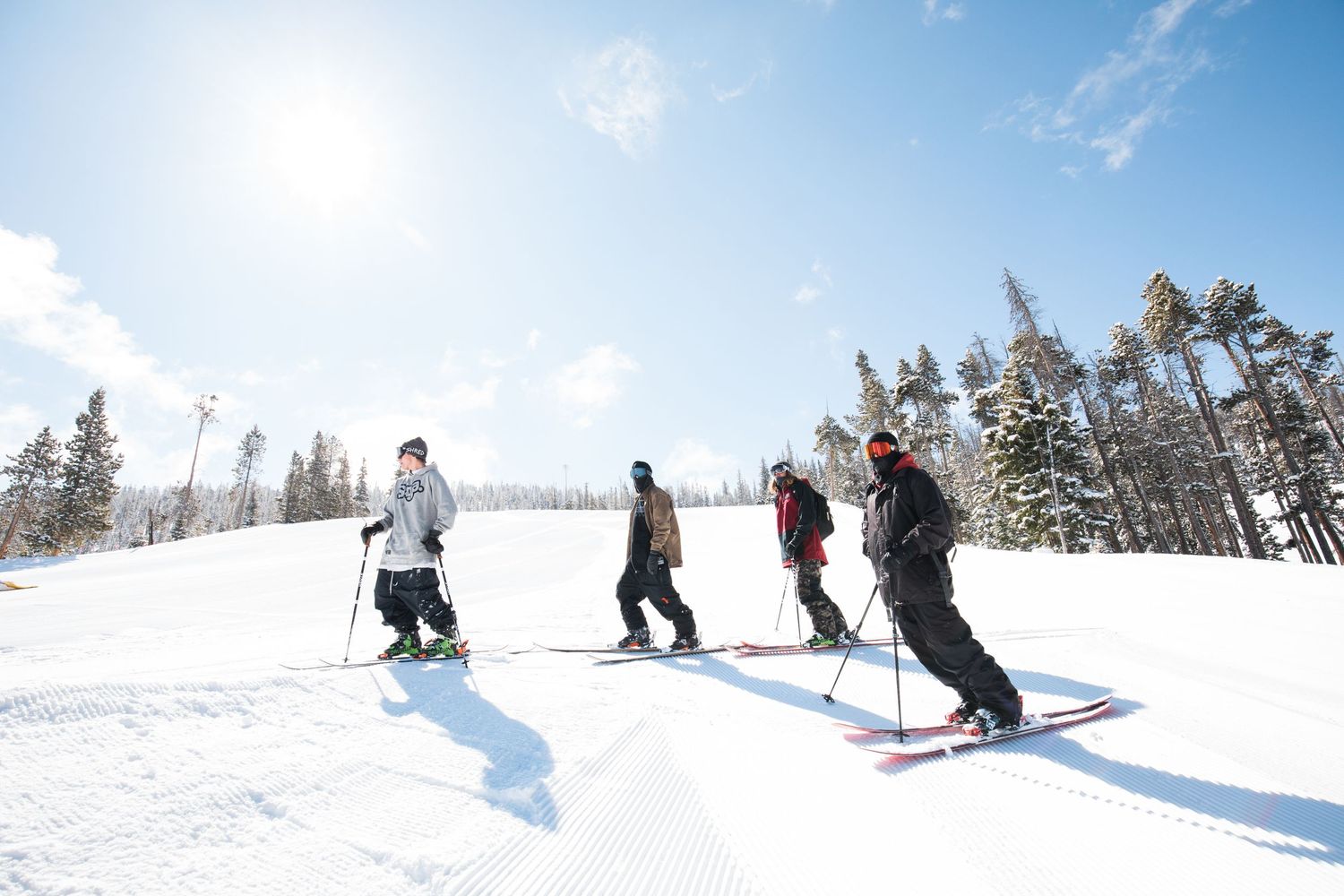 A group of skiers traverse across a snowy slope on a sunny winter day surrounded by evergreen trees.