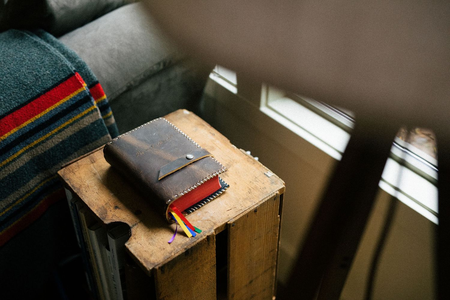 Leather-bound journal rests on rustic wooden surface near window with sunlight streaming through blinds.