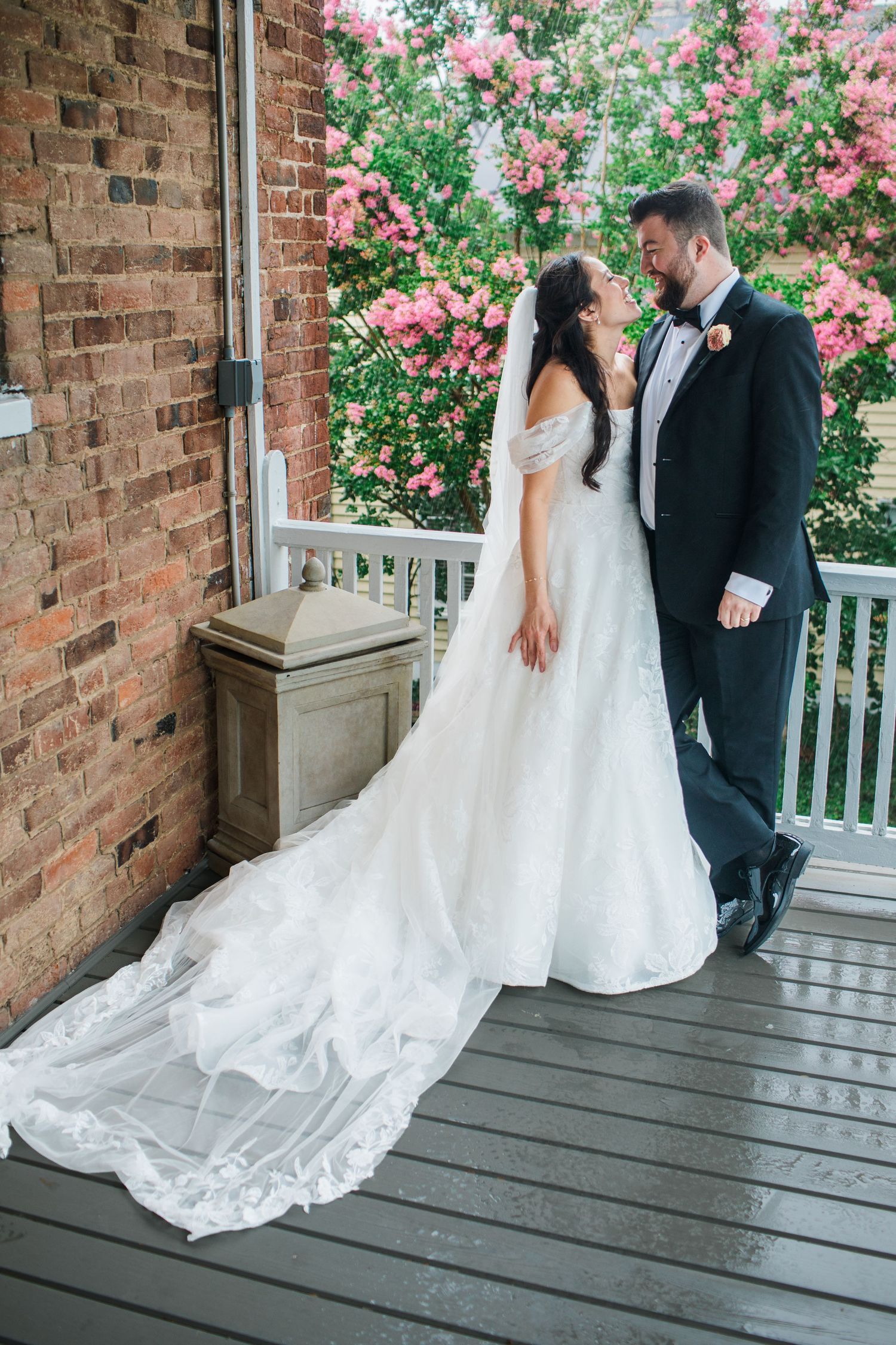 Bride and groom share a romantic moment on a wooden porch, surrounded by blooming pink flowers and brick walls.