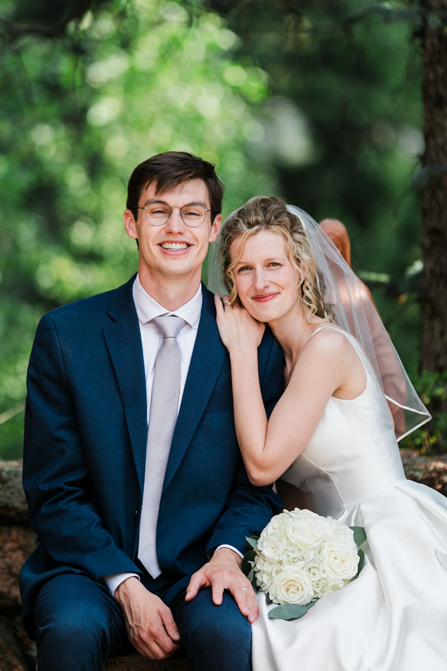 A smiling groom in a navy suit and bride in a white gown with veil holding white roses sit together outdoors.