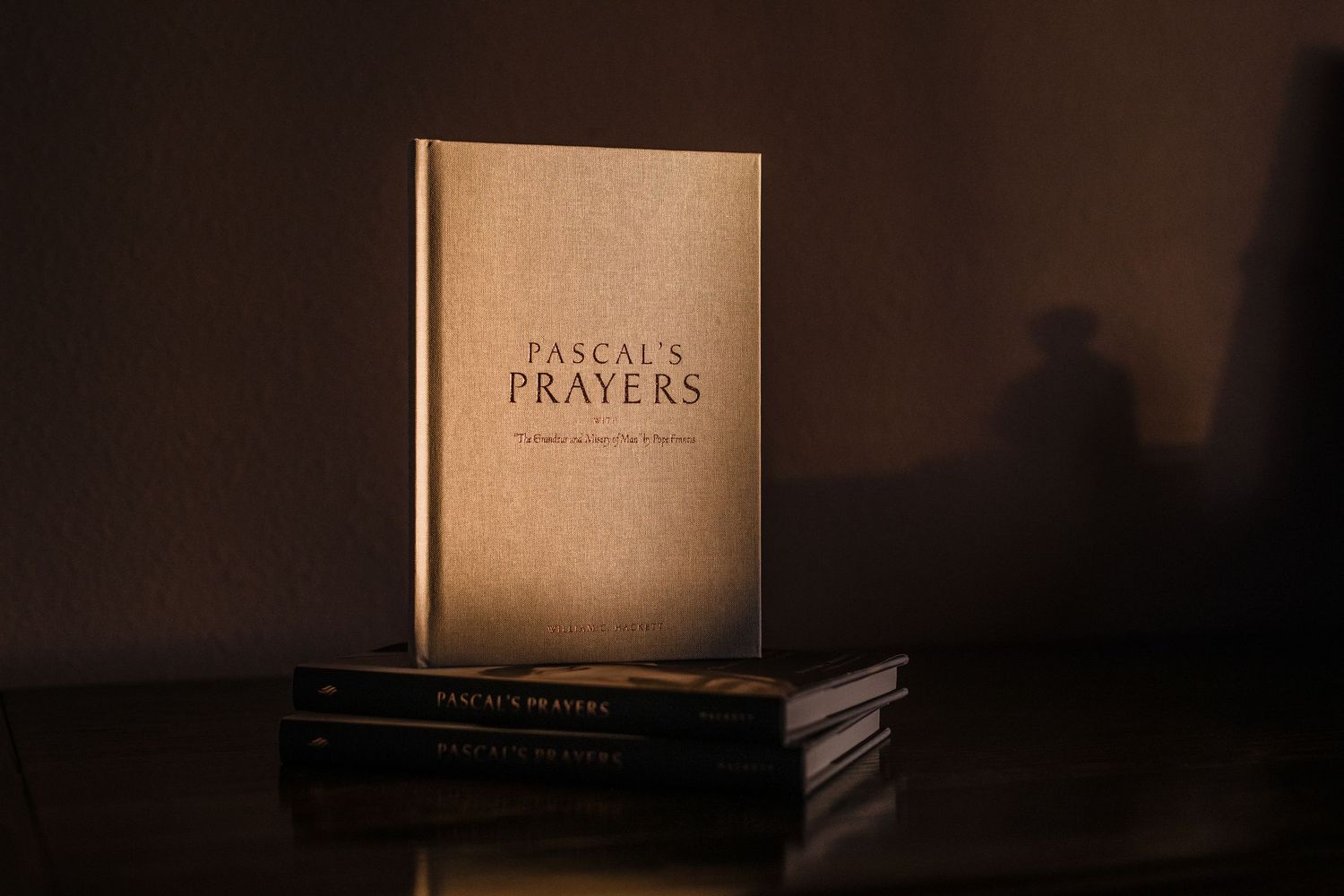 Gold hardcover edition of Pascal's Prayers book displayed upright on a dark reflective surface in moody lighting.