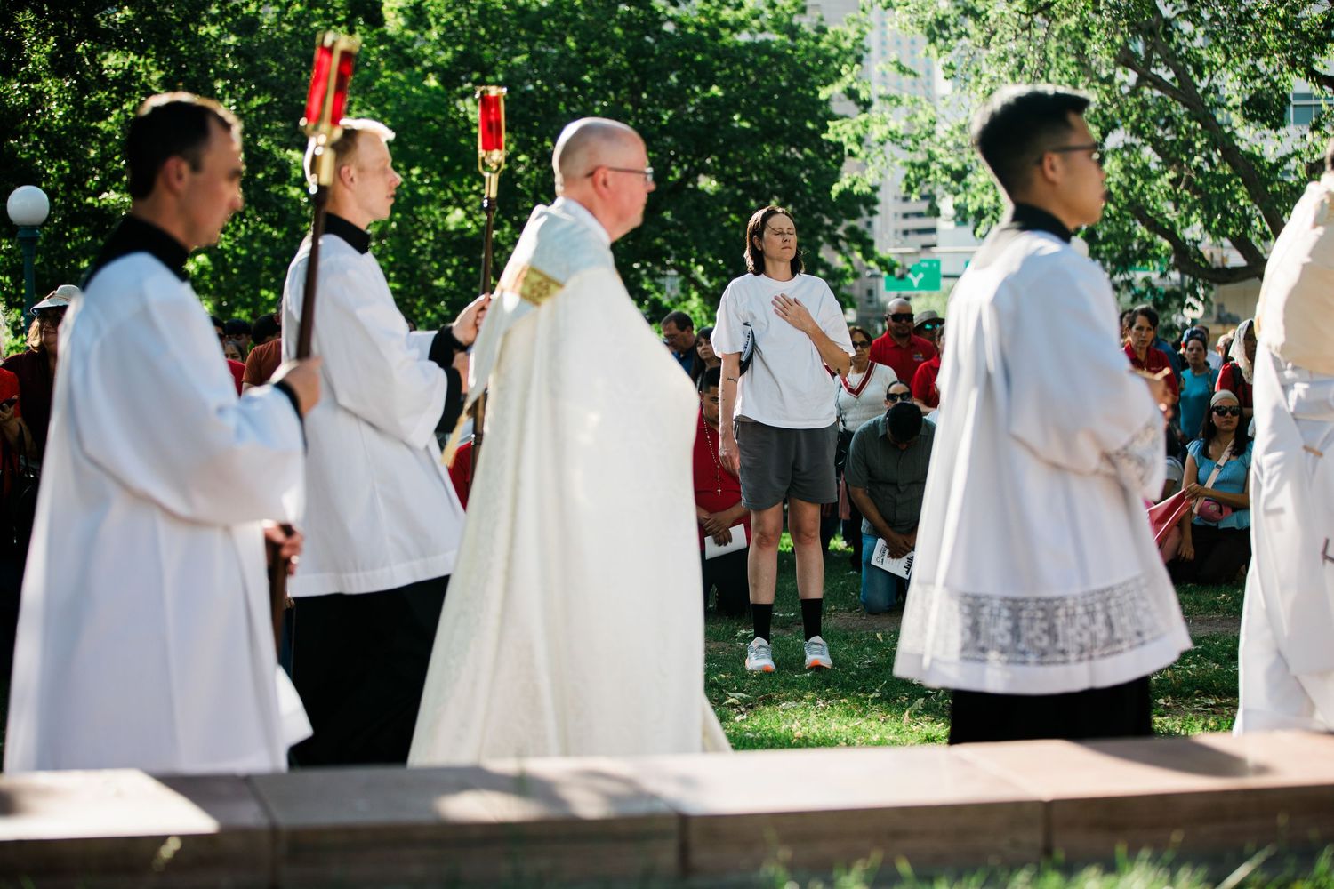 Catholic clergy in white vestments lead an outdoor religious procession surrounded by attendees in a sunny park setting.