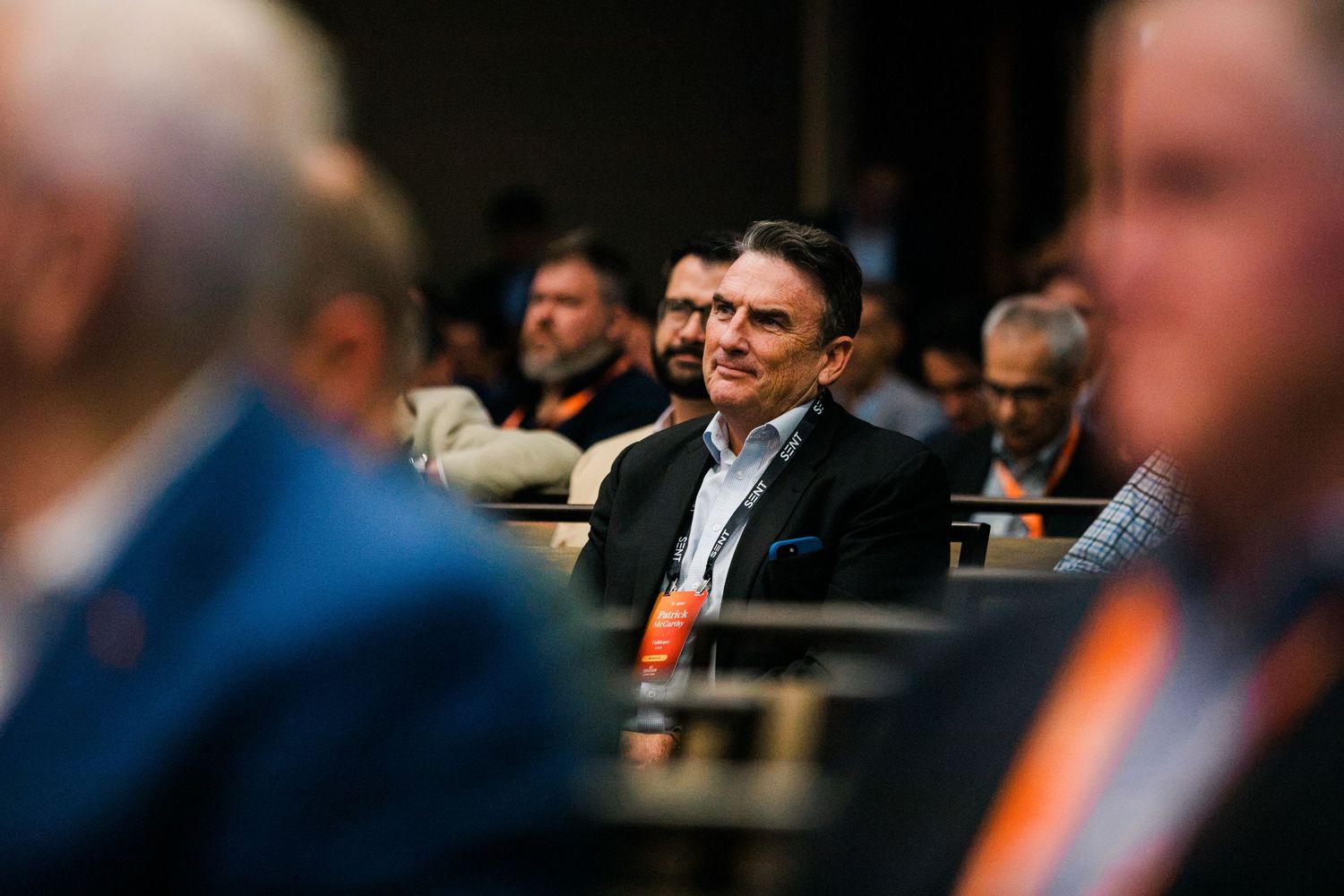 Man in blazer smiling at a conference event surrounded by attendees wearing orange lanyards.