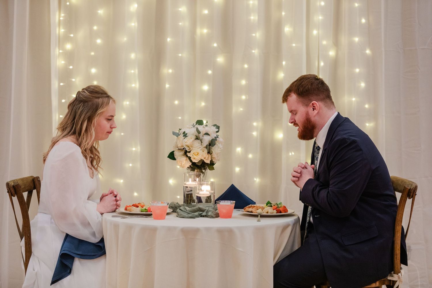 A couple shares an intimate dinner at a table decorated with flowers against a backdrop of twinkling string lights.