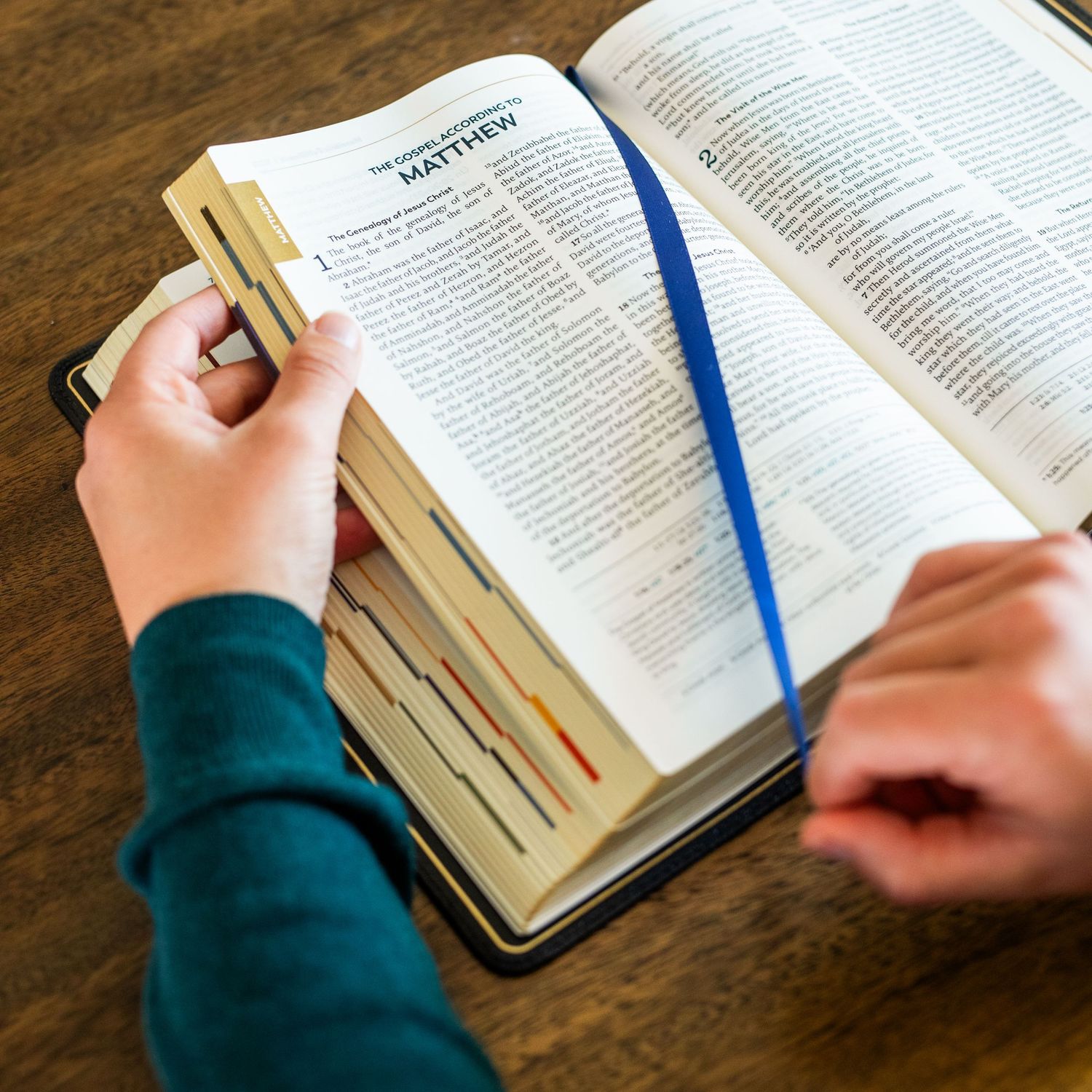 A person's hands are visible turning pages of an open Bible with a blue bookmark ribbon on a wooden table.