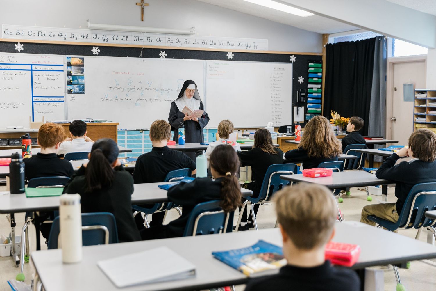 Students sit at desks while an instructor stands at the whiteboard in a bright classroom with posters on the walls.