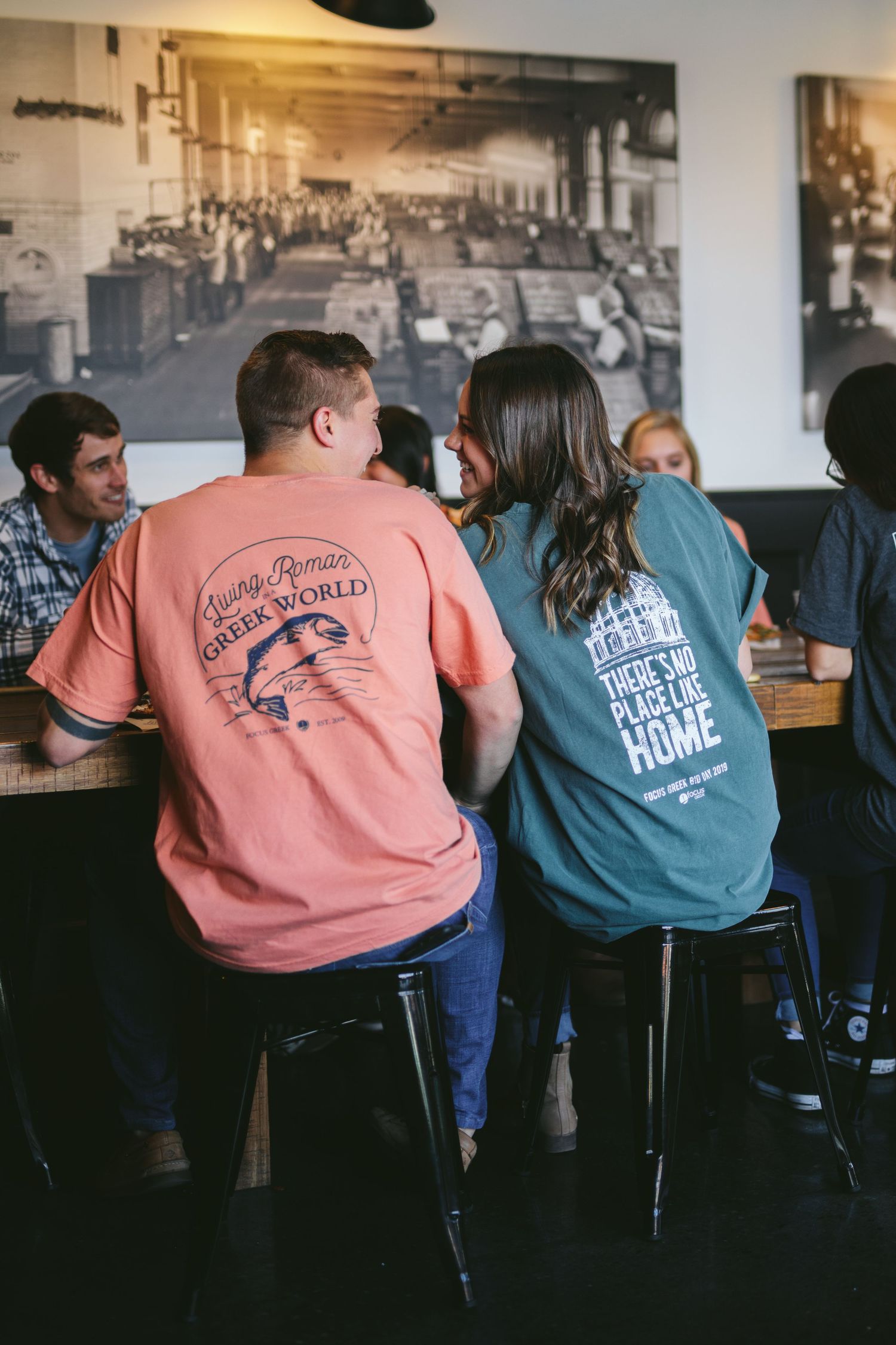 People wearing blue and coral colored t-shirts sit at a bar counter in a dimly lit establishment.