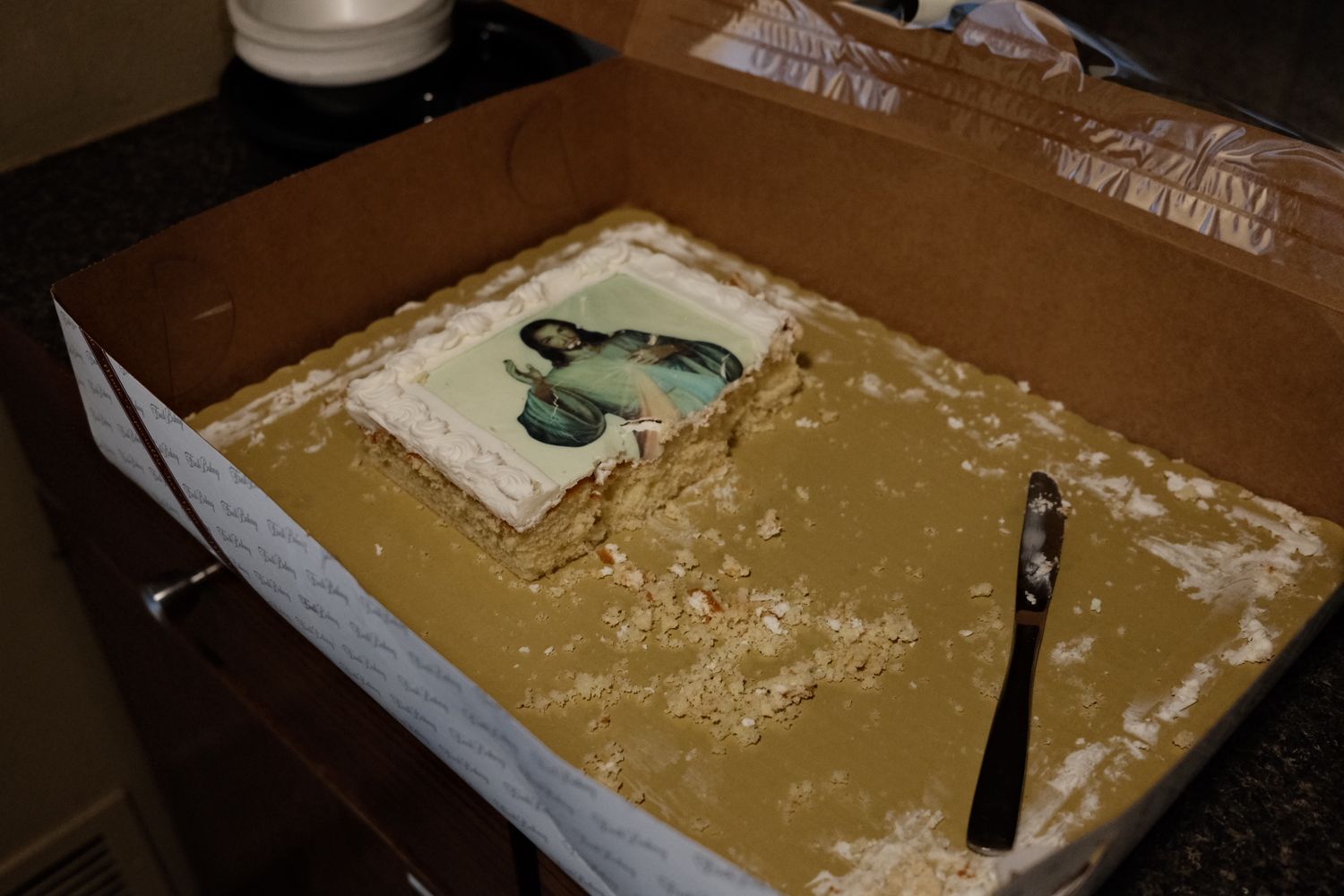 A messy cake with white frosting shows remnants of birthday celebration including a knife and green decorative piece.