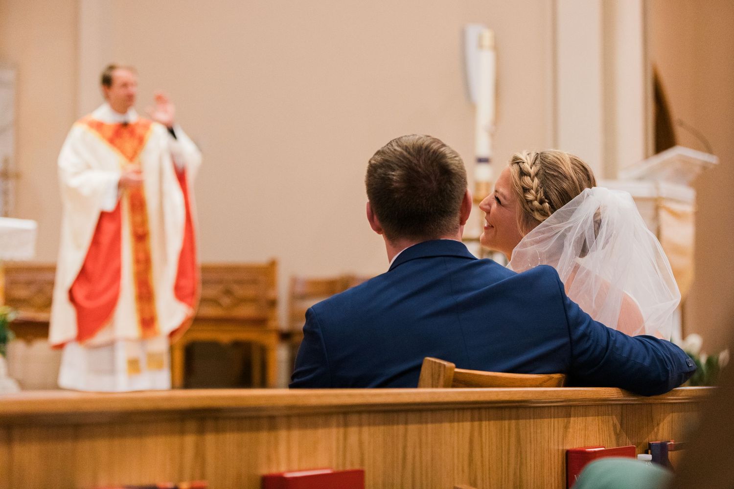 Bride and groom sit in church pew during wedding ceremony as priest in red vestments speaks at altar.