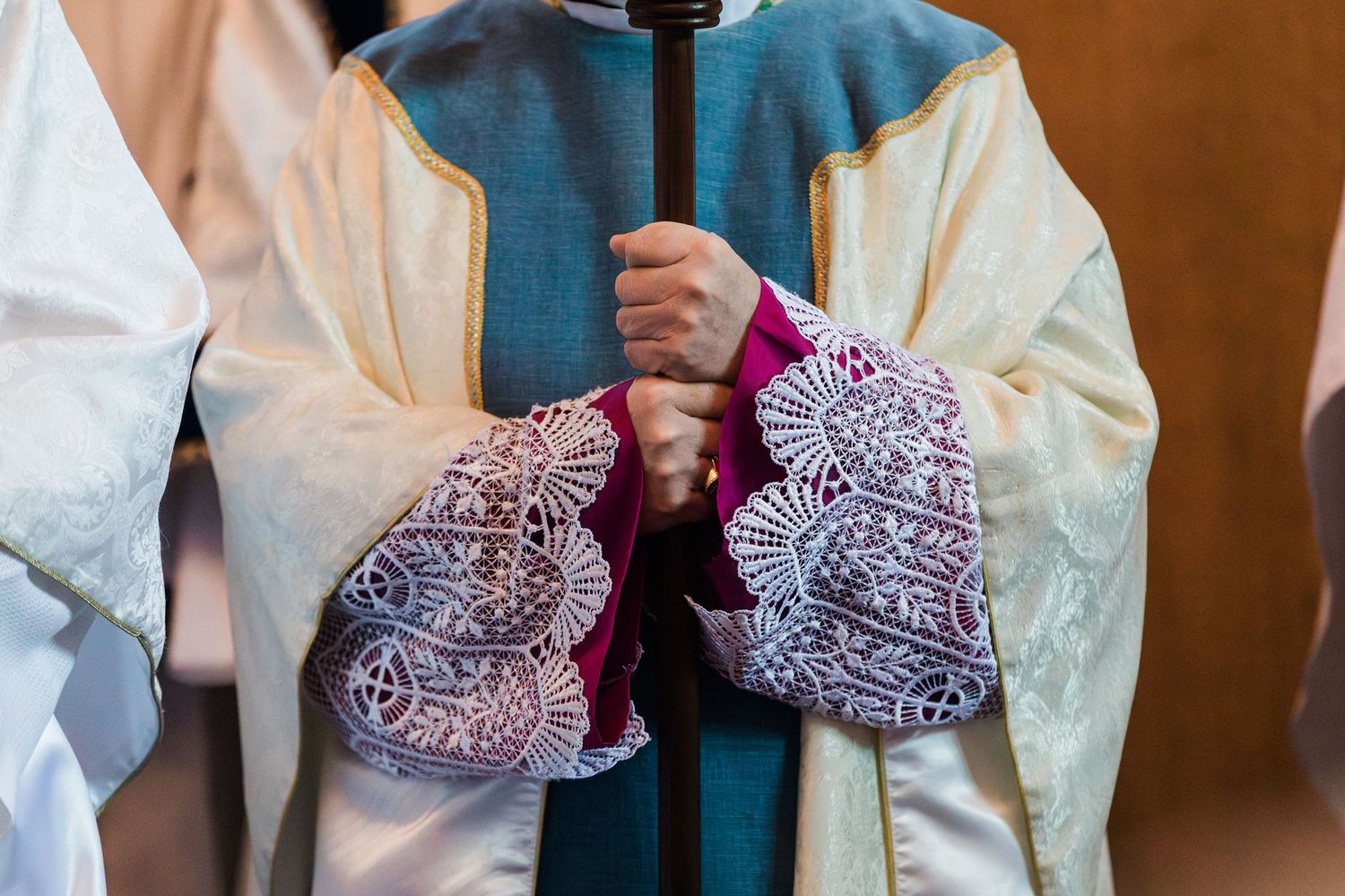 Close-up of Catholic clergy hands holding a ceremonial staff wearing ornate embroidered liturgical vestments.